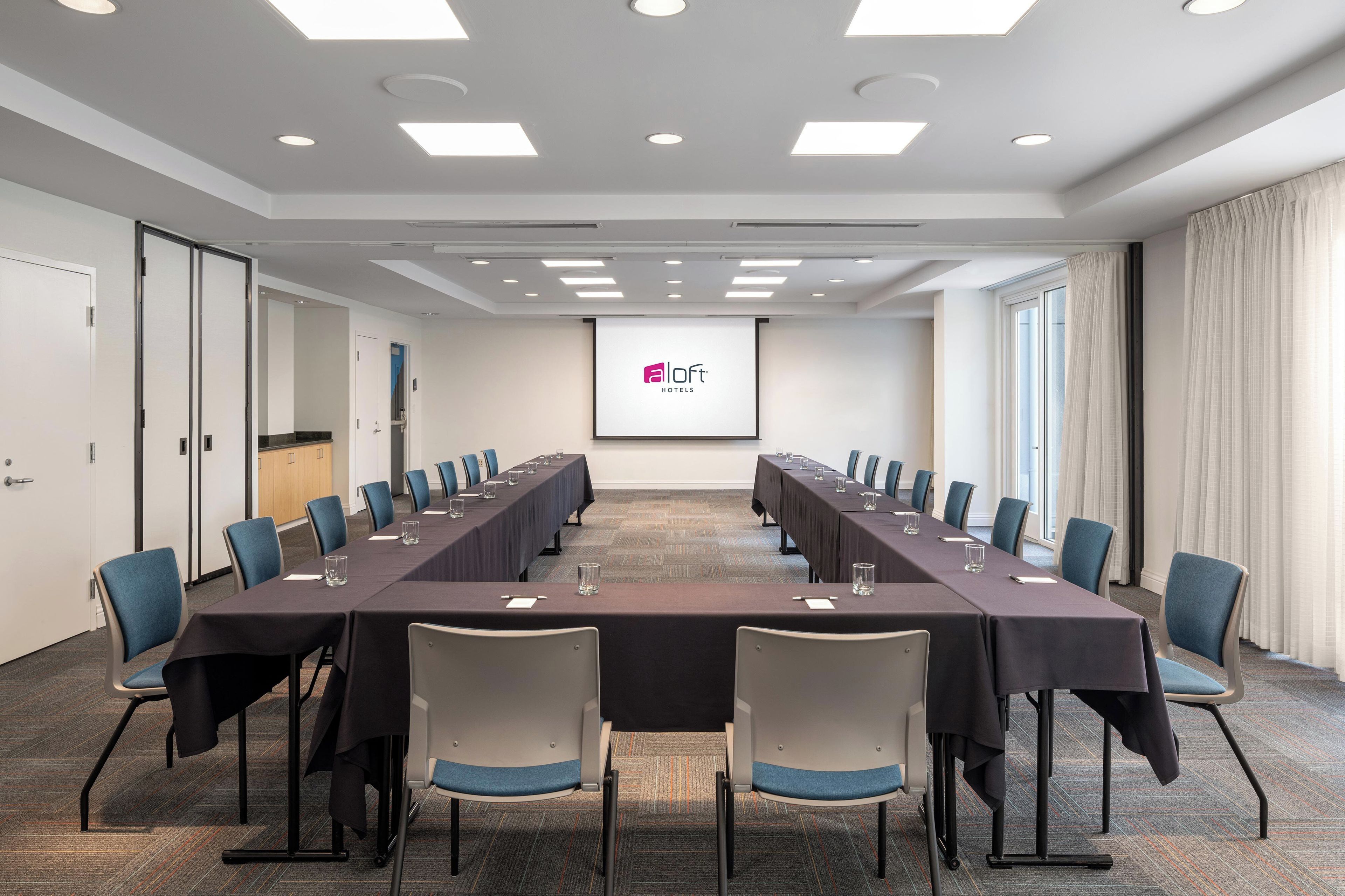 Un salón de conferencias con una larga mesa, sillas y una pantalla mostrando un logotipo.