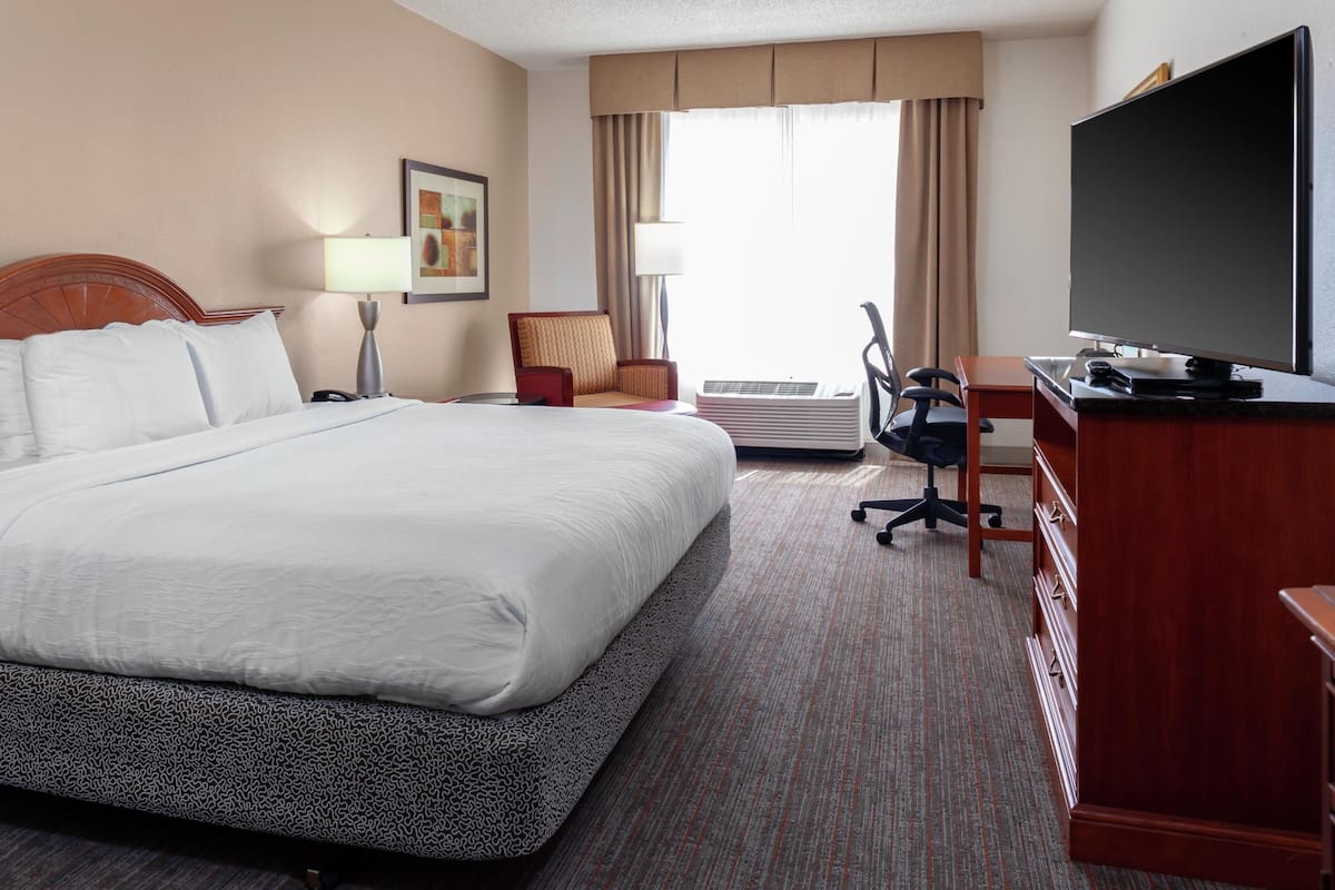A hotel room with a large bed, a desk with a television, an air conditioning unit, and a chair.