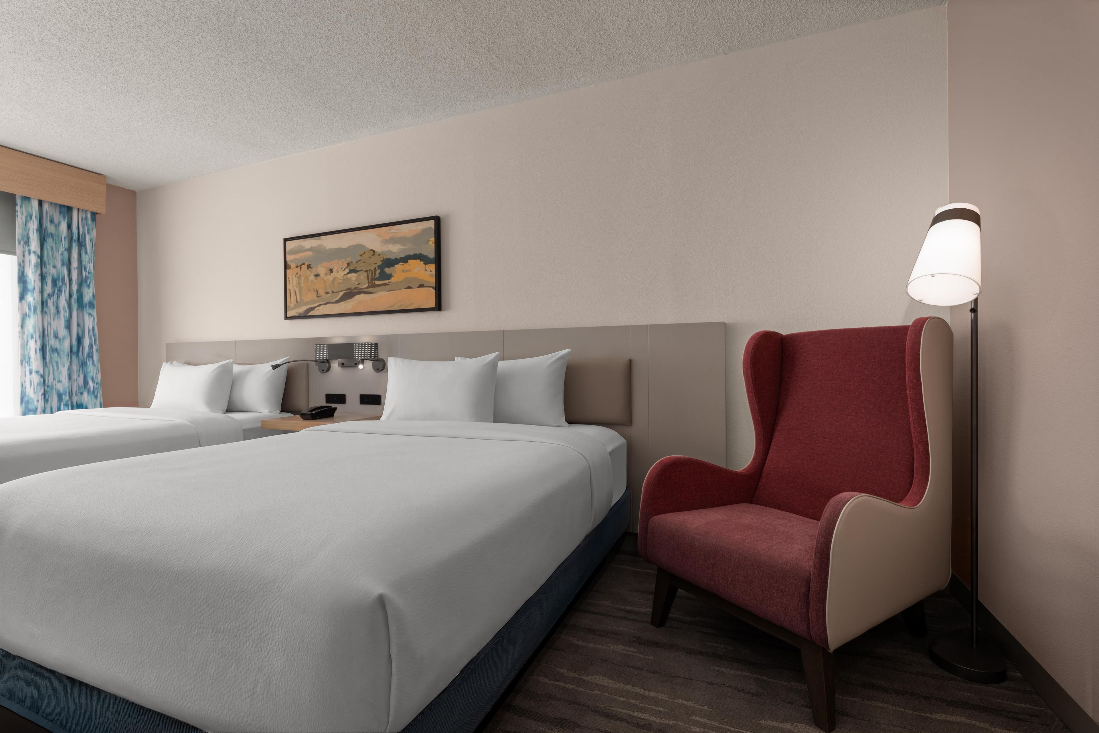 Habitación de hotel con dos camas, un sillón rojo, una lámpara y un cuadro en la pared.