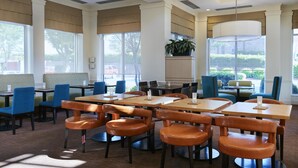A spacious dining area with multiple tables and chairs, large windows with blinds, and a potted plant.