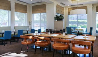 A spacious dining area with multiple tables and chairs, large windows with blinds, and a potted plant.