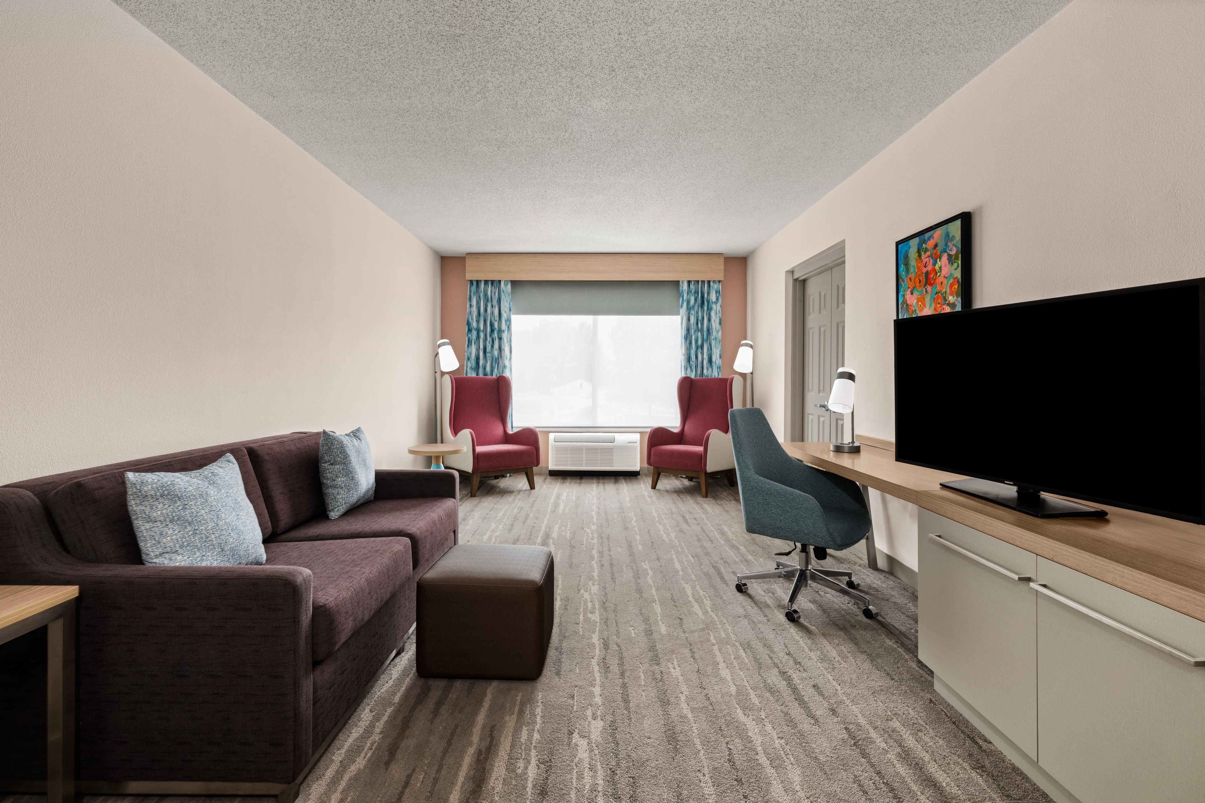 A hotel room with a sofa, two armchairs, a desk with a flat-screen TV, and a large window with curtains.