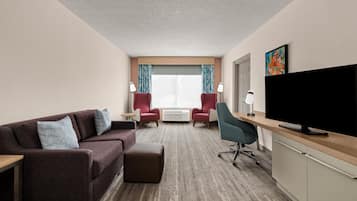 Une chambre d’hôtel avec un canapé, deux fauteuils, un bureau avec un téléviseur à écran plat et une grande fenêtre avec des rideaux.