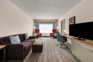 Quarto de hotel com sofá, duas poltronas, escrivaninha e televisão de tela plana, além de uma janela grande com cortinas.