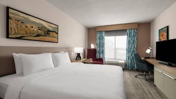 A hotel room with a large bed, a desk with a flat-screen TV, a chair, a desk lamp, a window with curtains, and a painting on the wall.