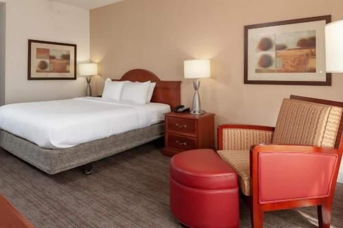 A hotel room with a bed, a nightstand with a lamp, a chair, and a red ottoman.