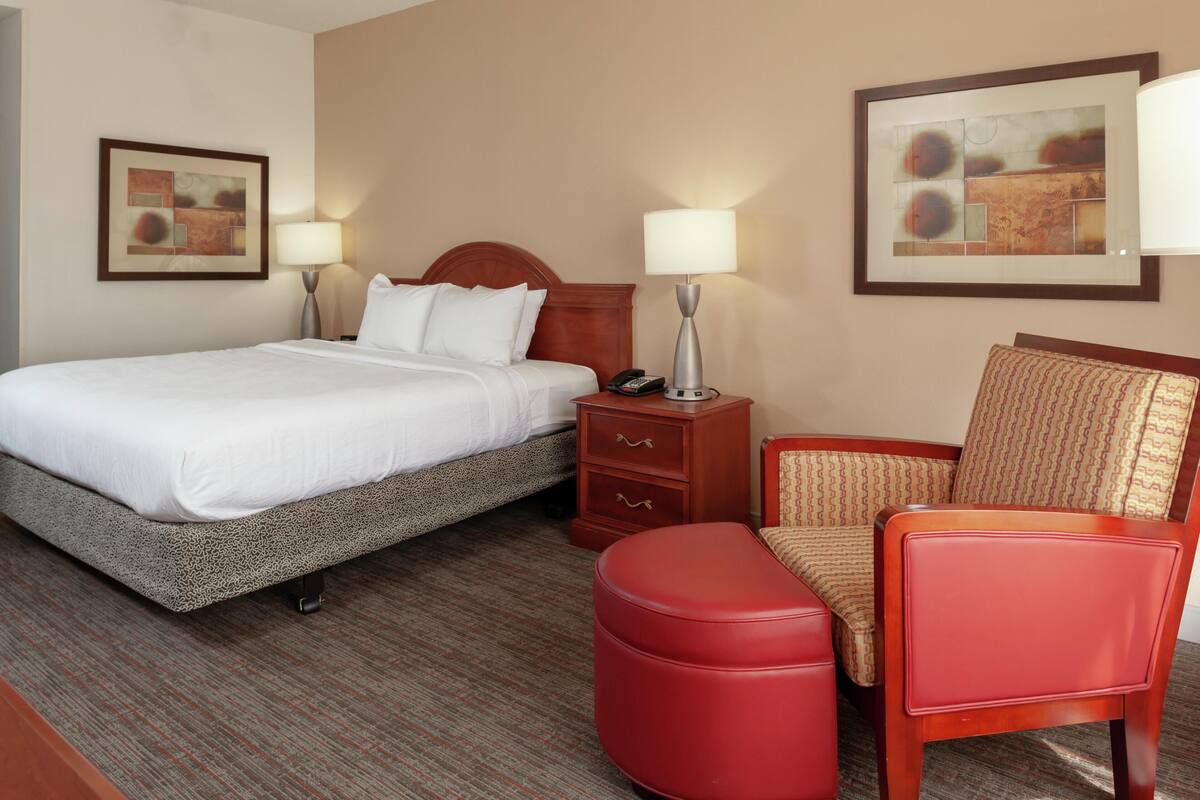 A hotel room with a bed, a nightstand with a lamp, a chair, and a red ottoman.