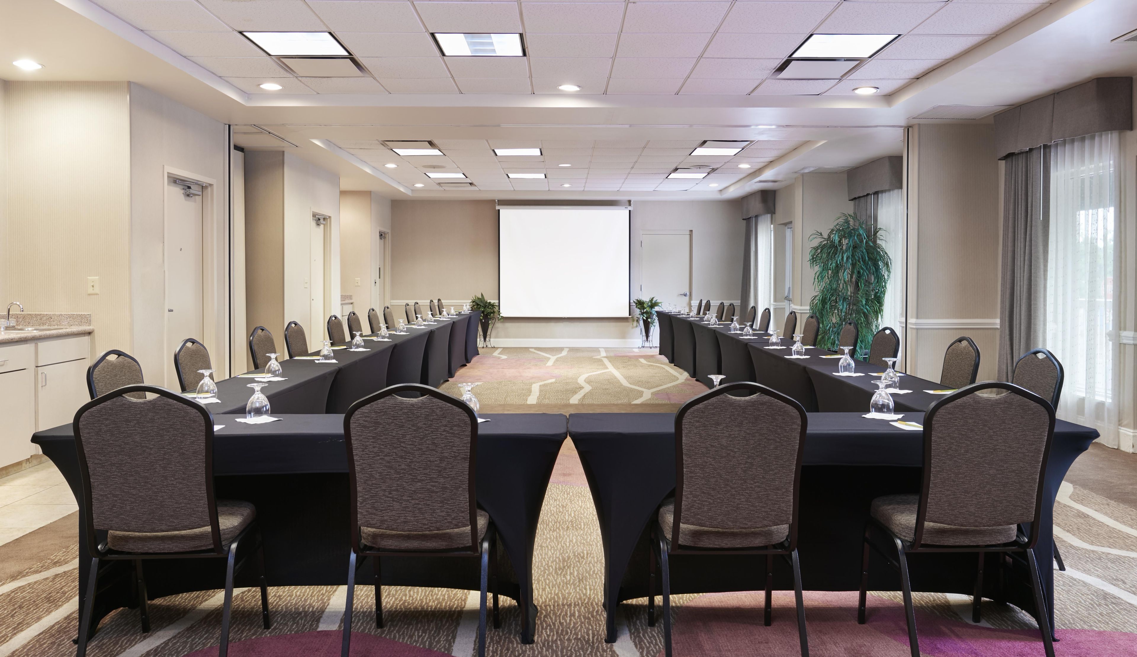 A conference room with a long table, chairs, a projector screen, and a plant.