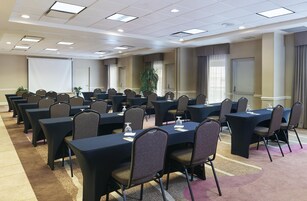 Un salón de conferencias con varias mesas y sillas, una pantalla de proyector y un ventanal con cortinas.