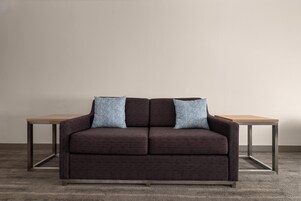 Un canapé brun foncé, agrémenté de deux coussins assortis, est placé contre un mur uni, et une table en bois se trouve de chaque côté.
