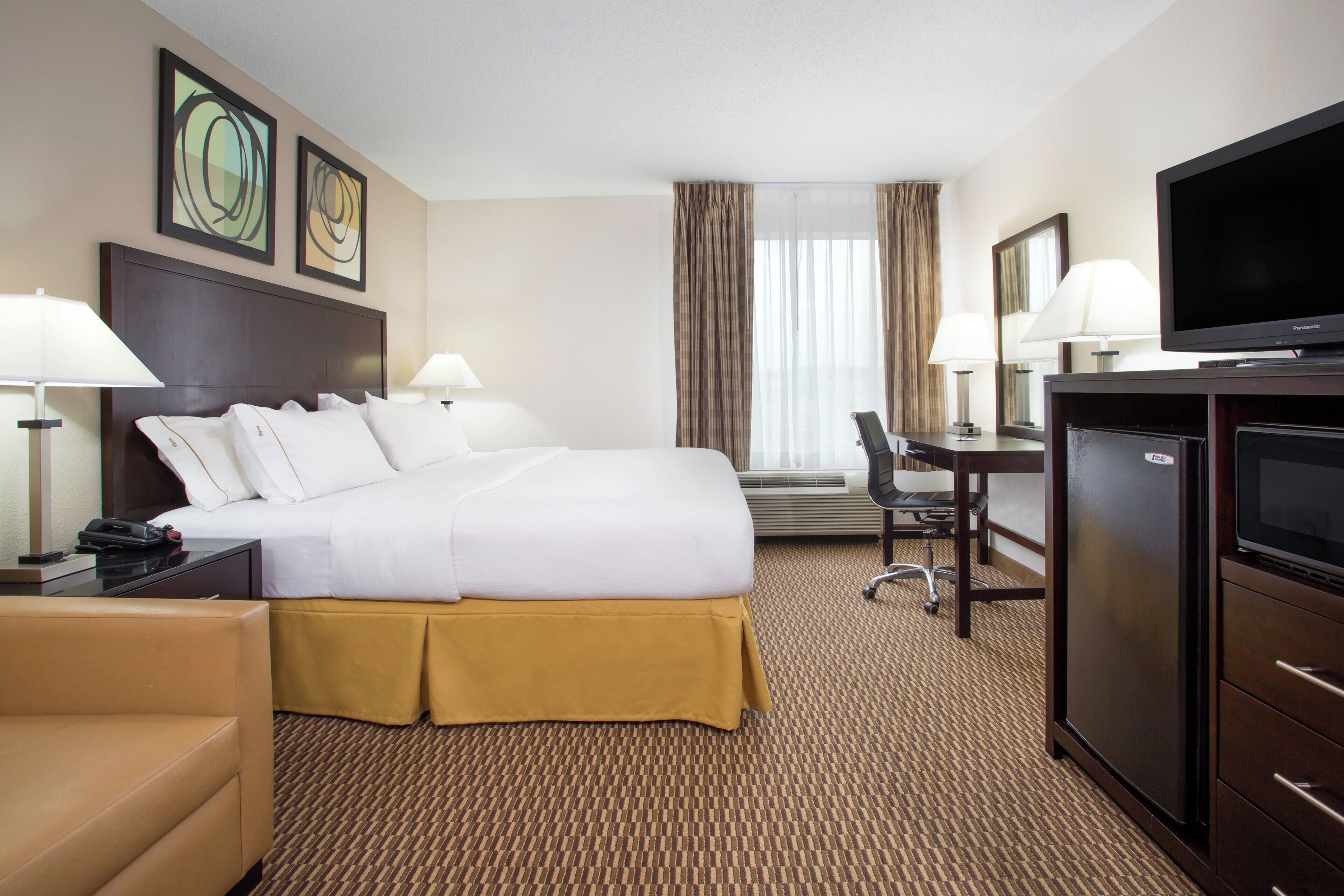 Une chambre d’hôtel comprenant un lit, des lampes de chevet, un bureau avec une chaise, une télévision et une grande fenêtre avec des rideaux.
