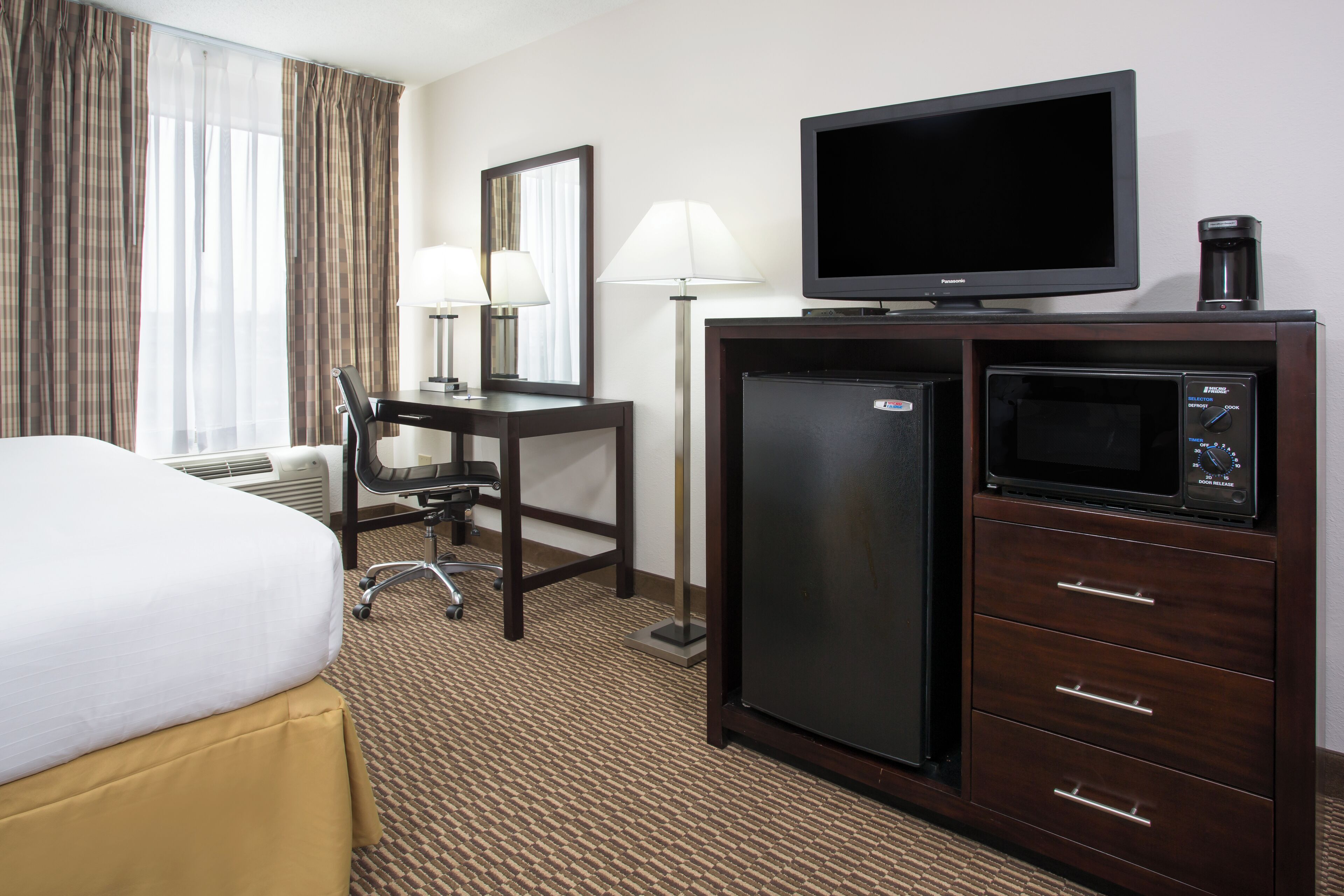 A hotel room with a bed, desk, chair, lamp, television, refrigerator, and microwave.
