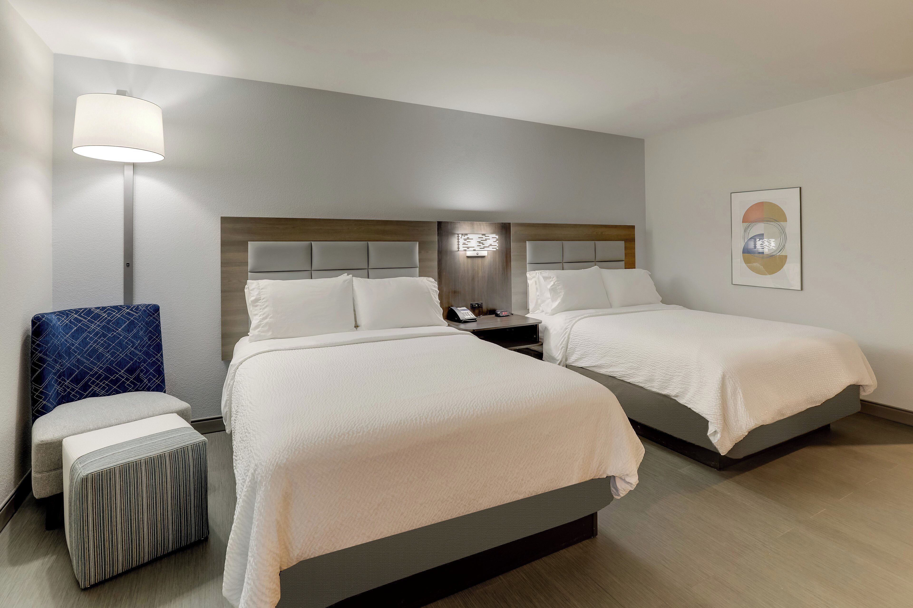 A hotel room with two beds, a nightstand, a lamp, a chair, and a small table.