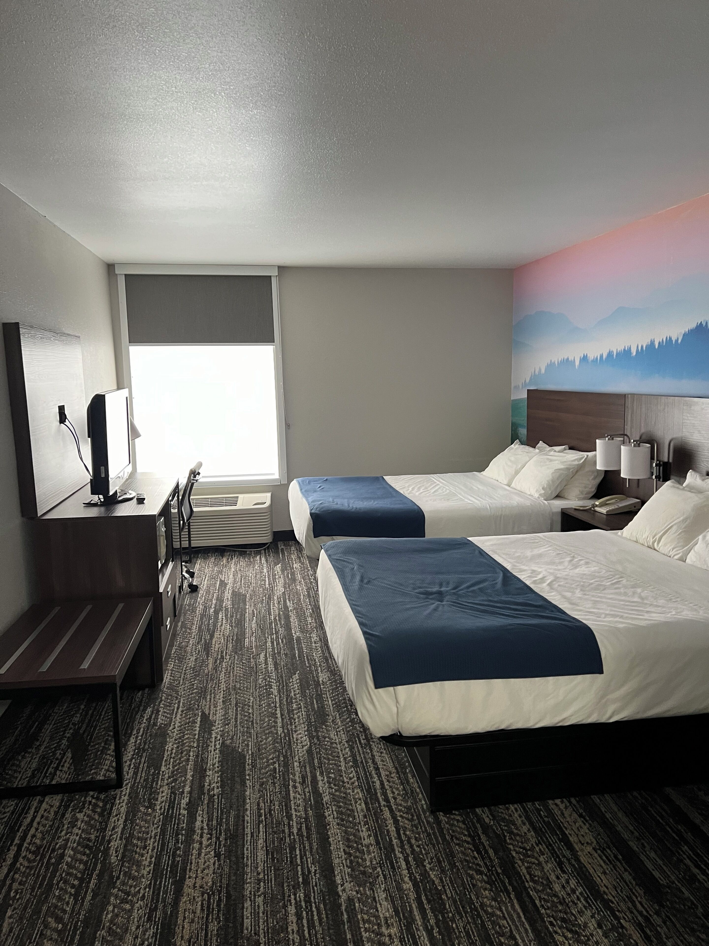 A hotel room with two beds, a desk, a chair, a TV, and a painting on the wall.