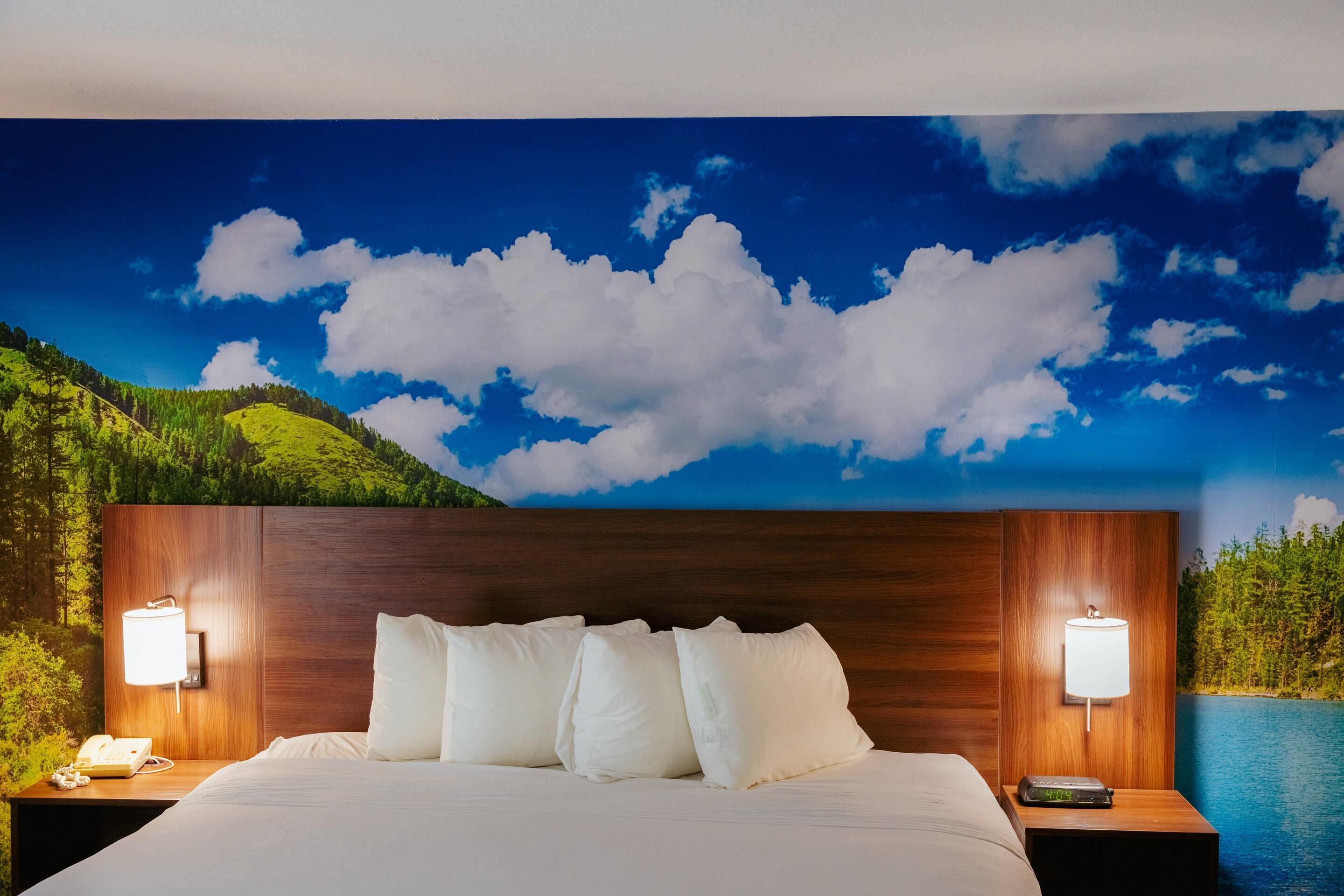 Une chambre d’hôtel avec un lit, deux tables de chevet avec des lampes, une fresque murale représentant un paysage et une vue sur une étendue d’eau.