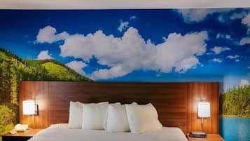 Habitación de hotel con cama, dos mesitas de noche con lámparas, un mural de paisaje en la pared y vista a una masa de agua.