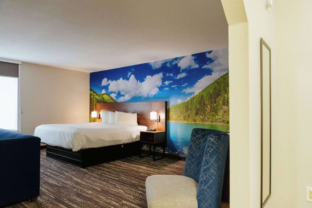 A hotel room with a bed, a desk, a chair, and a large mural of a mountain landscape.