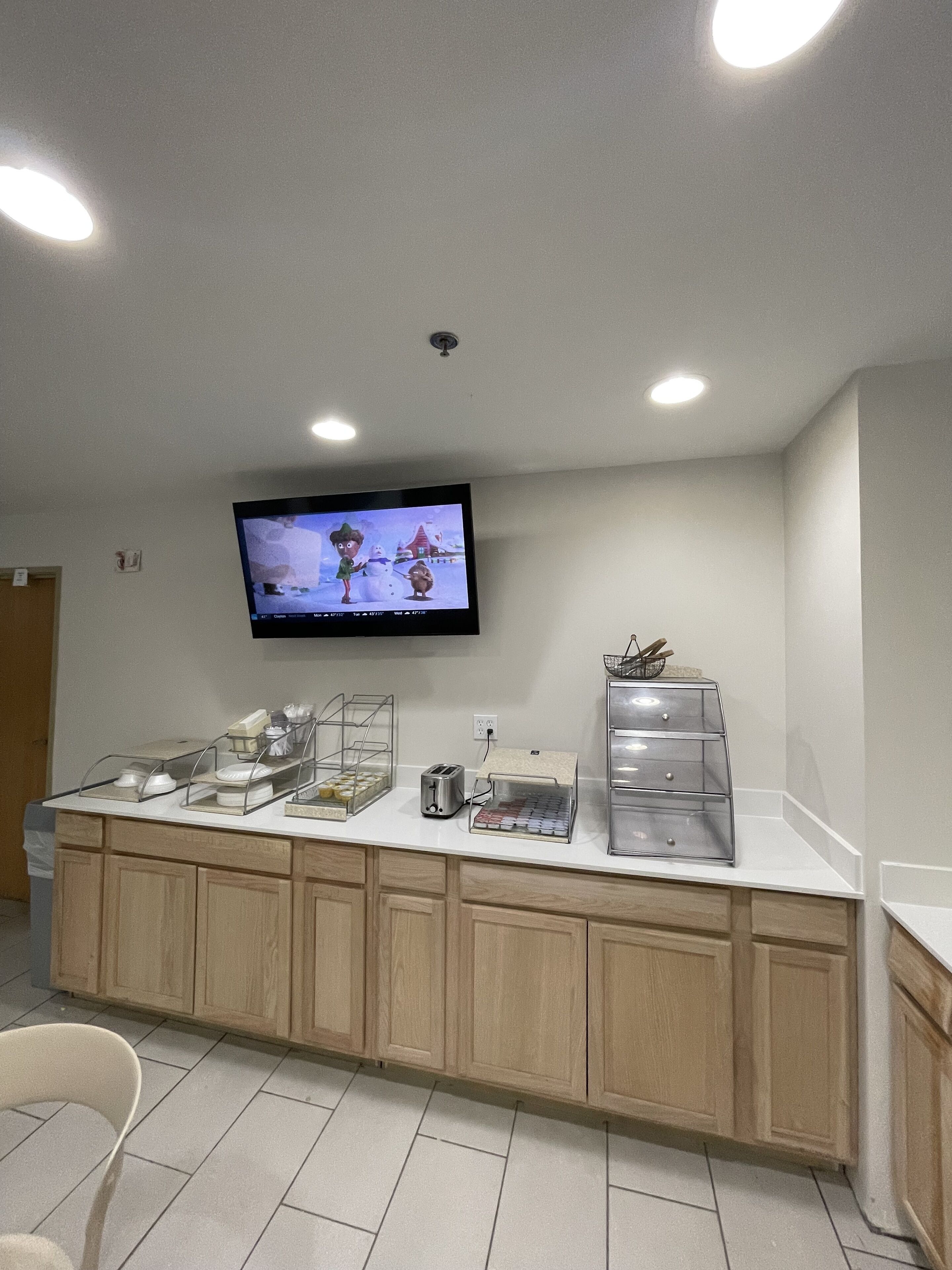 A clean, modern kitchen area with a flat-screen TV mounted on the wall, a buffet counter with various food items, and wooden cabinets.