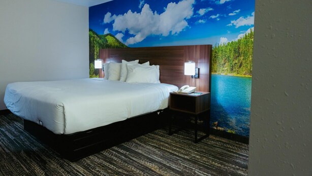 A hotel room with a large bed, a nightstand with a lamp, and a mural of a lake and mountains on the wall. - Clarion Pointe-Dillard near Blue Ridge Mountains (Dillard)