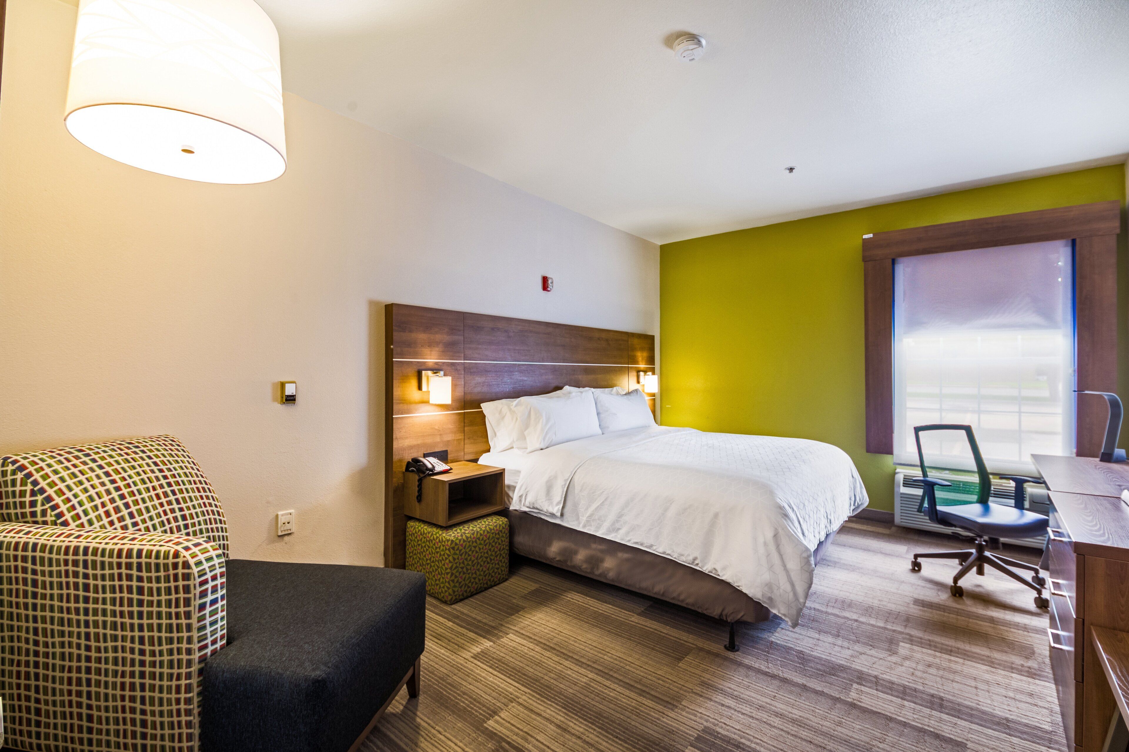 A hotel room with a bed, a desk, a chair, a nightstand, and a lamp.