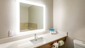 A bathroom with a white sink, a mirror, and a toilet. There are toiletries on the sink and a towel dispenser. - Holiday Inn Express Hotel & Suites Columbus by IHG (Columbus)