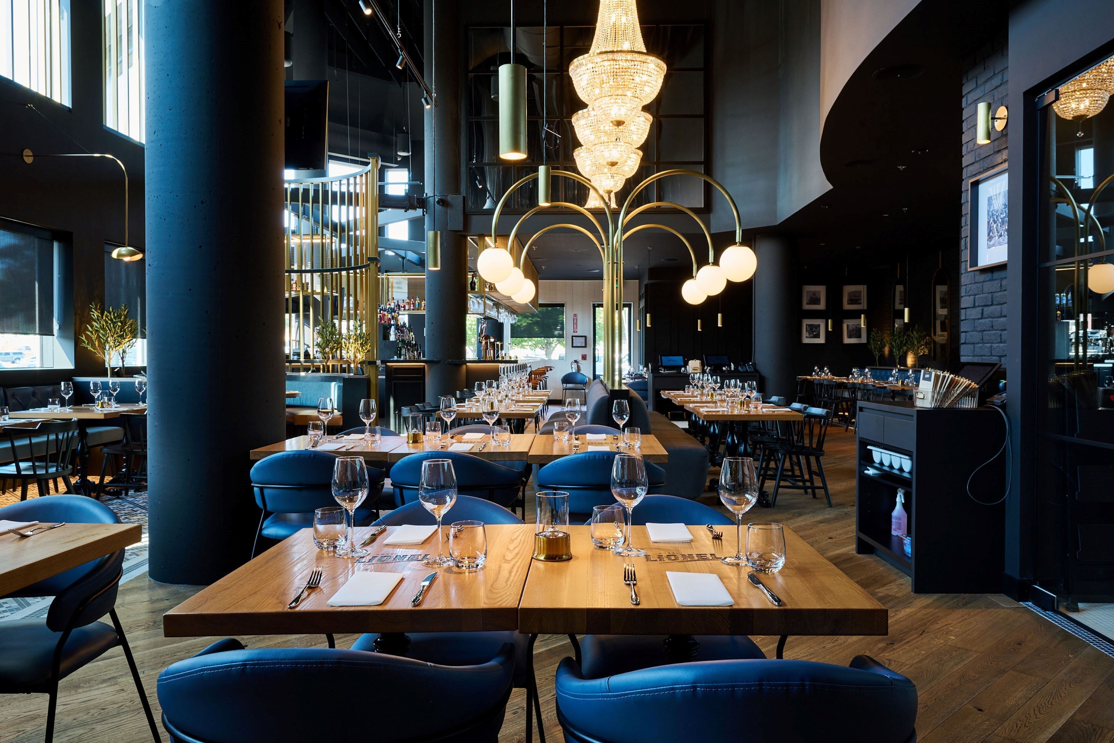Un restaurant doté de nombreuses tables, de chaises recouvertes d’un tissu bleu et d’un lustre central.