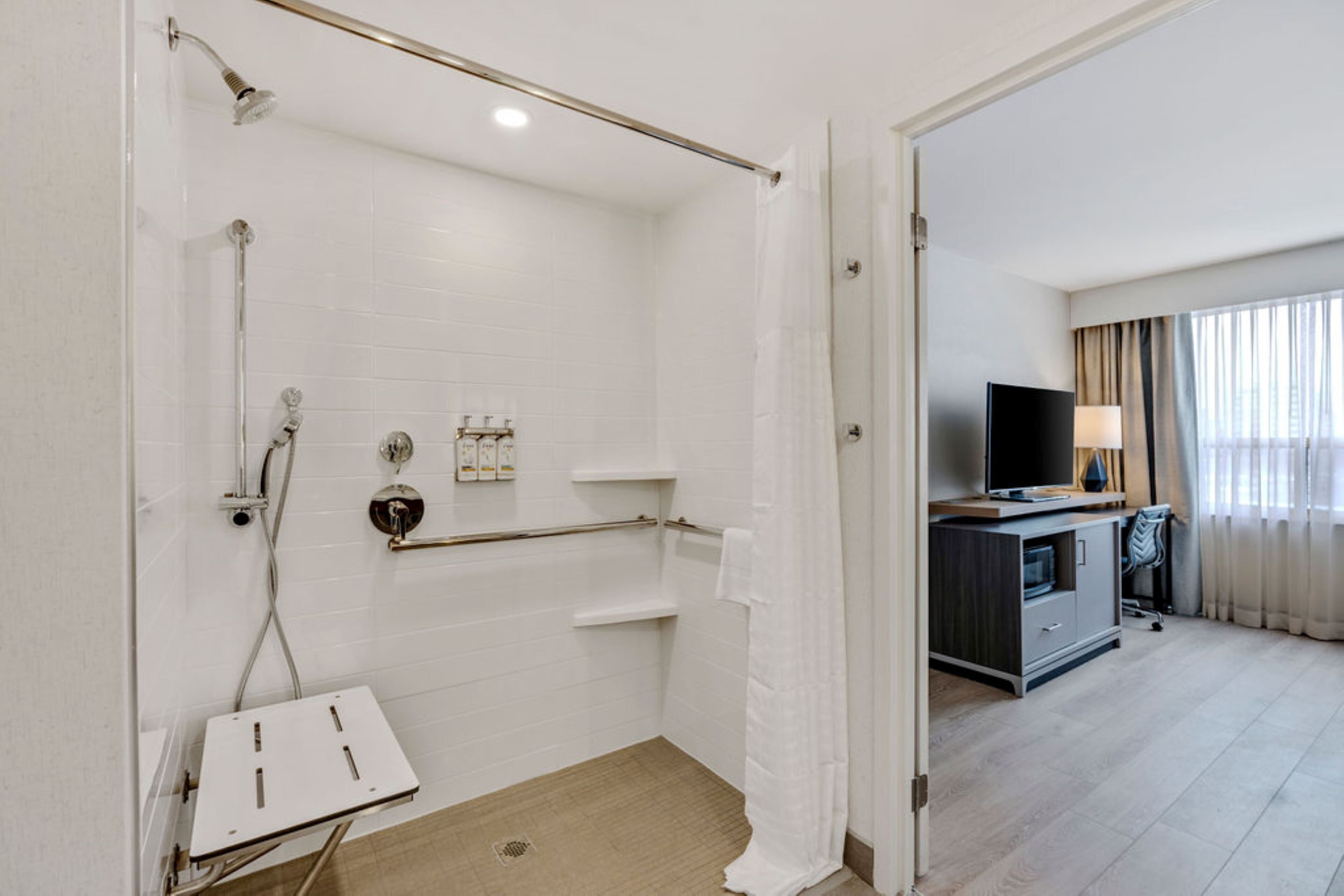 A hotel bathroom with a shower, toilet, and sink, a television, and a desk with a chair.