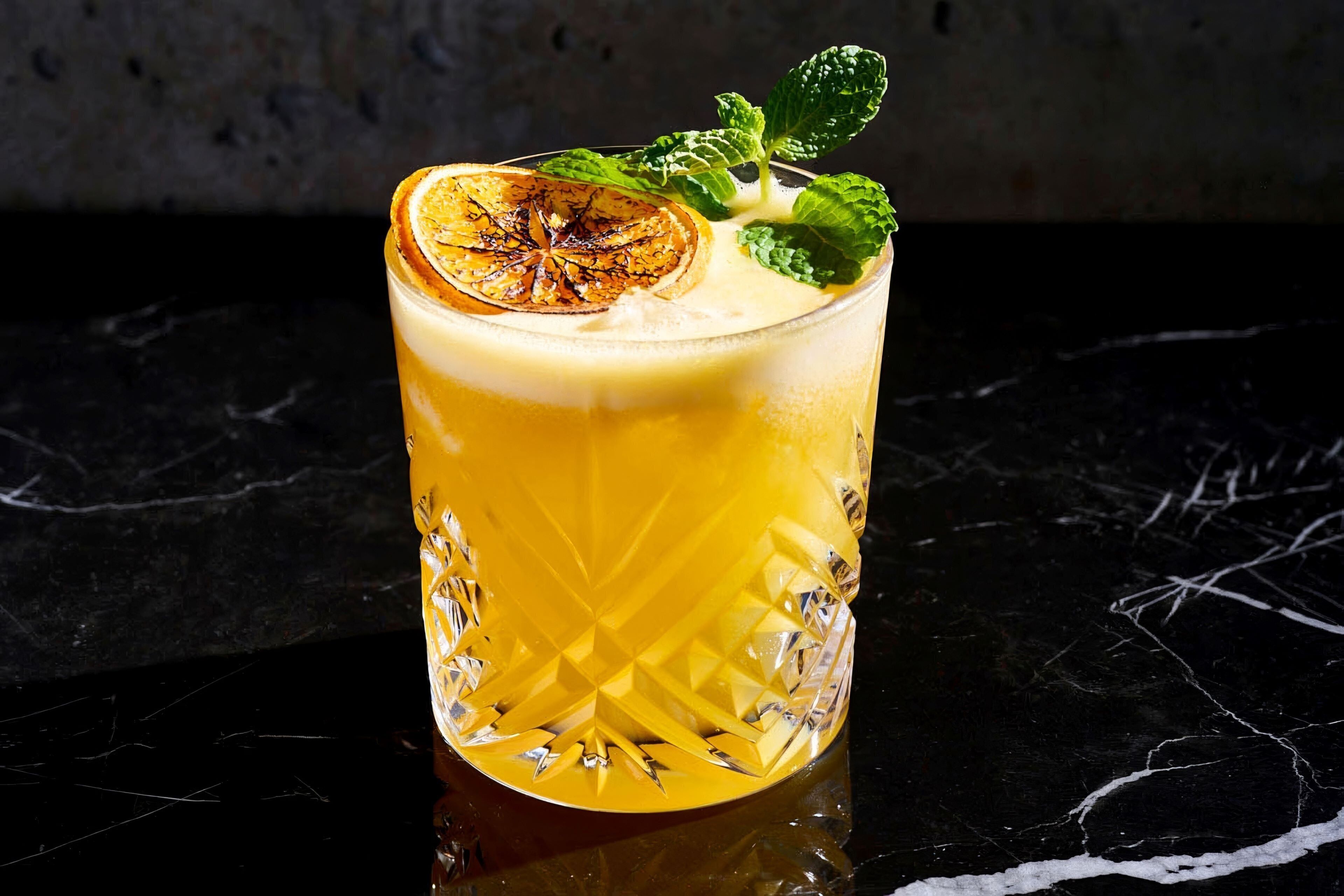 Un vaso de jugo de naranja con una rodaja de naranja y hojas de menta sobre una superficie de mármol.