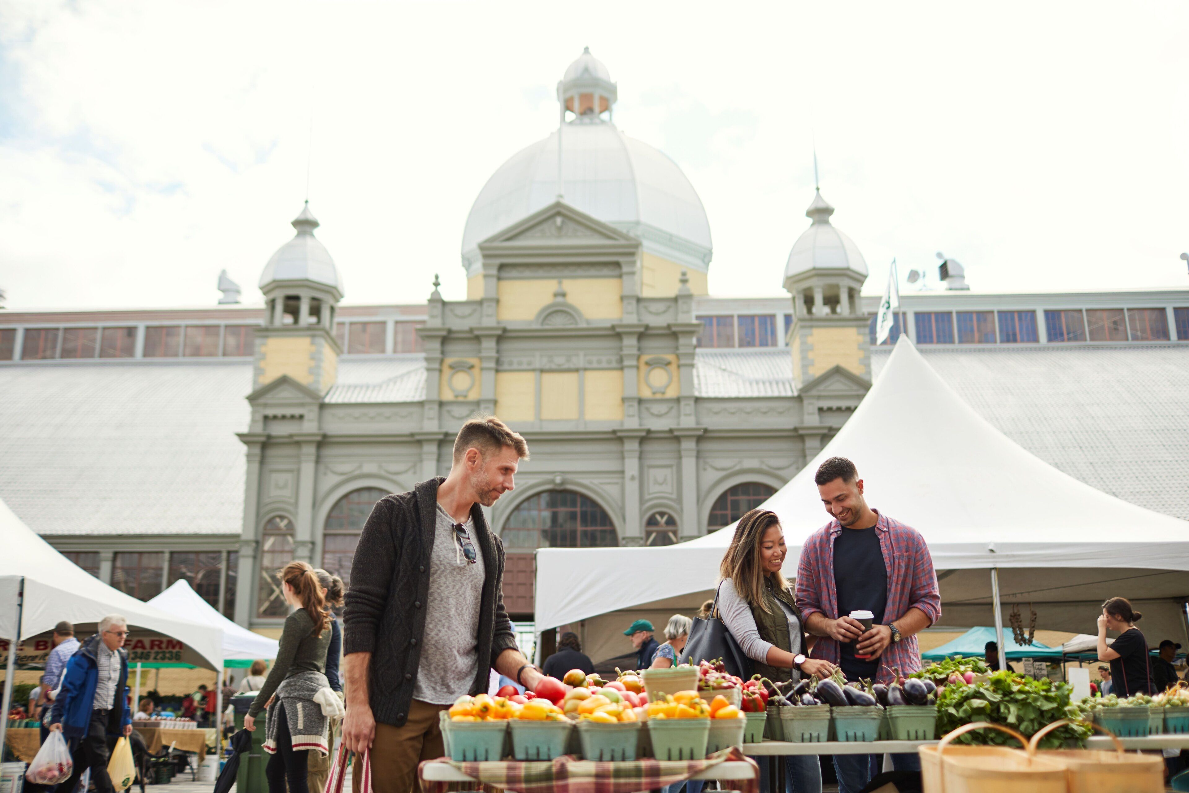 A market with people browsing stalls, set against a historic building with domed architecture.
