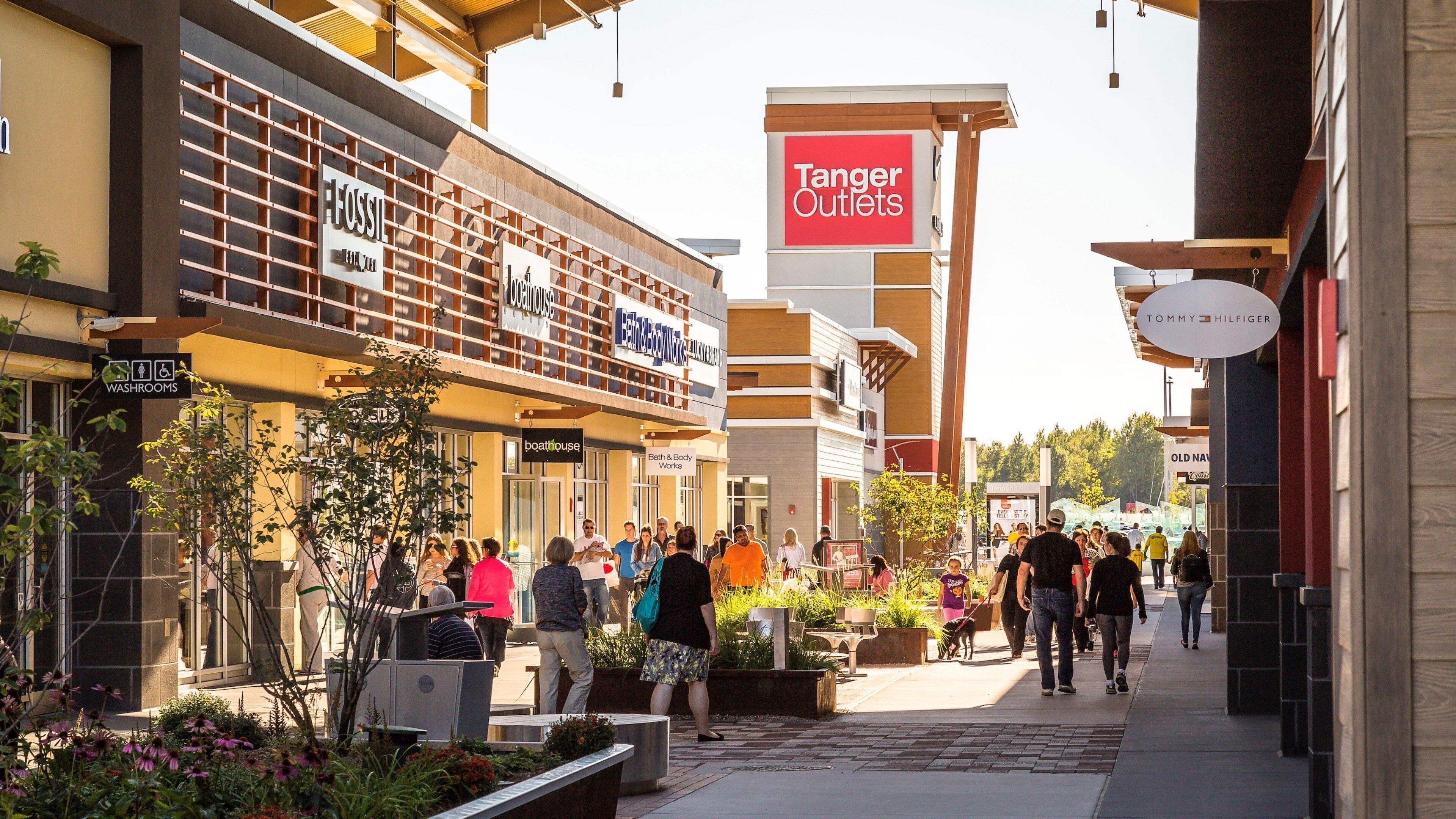 Eine Einkaufsstraße mit verschiedenen Geschäften, darunter ein Schild von Tanger Outlets, ein Bath & Body Works und ein Tommy Hilfiger Geschäft.