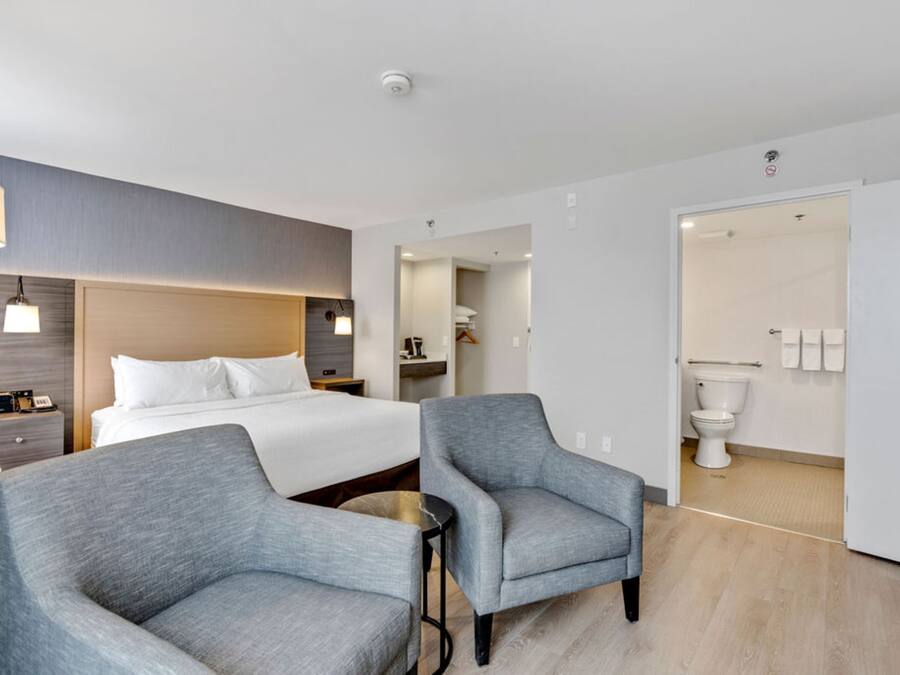 Una habitación de hotel moderna con una cama, dos sillones, una mesita, una lámpara de noche, un buró y un baño visible a través de una puerta abierta.