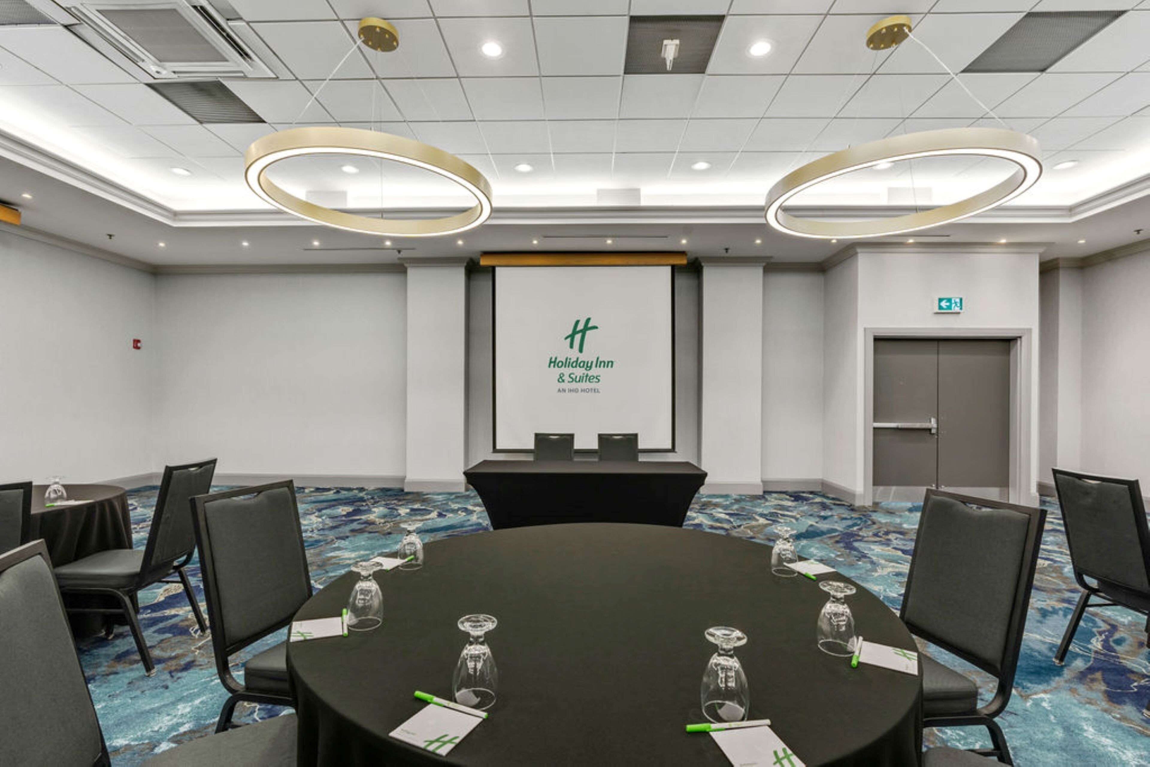 Una sala de conferencias con una mesa redonda, sillas negras y una pantalla mostrando el logotipo de Holiday Inn.