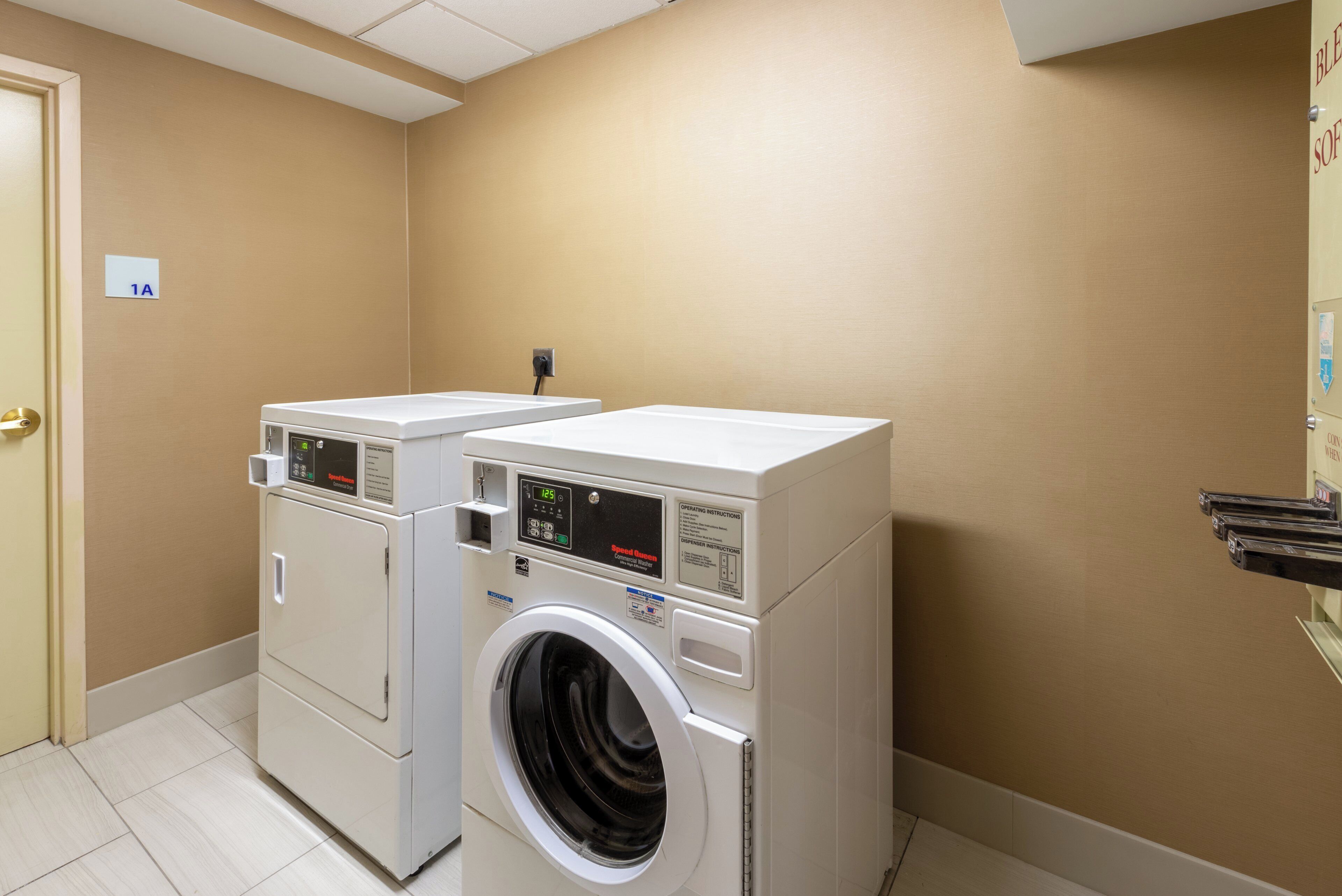 A laundry room with two stacked washers and dryers, a beige wall, and a door marked "1A".