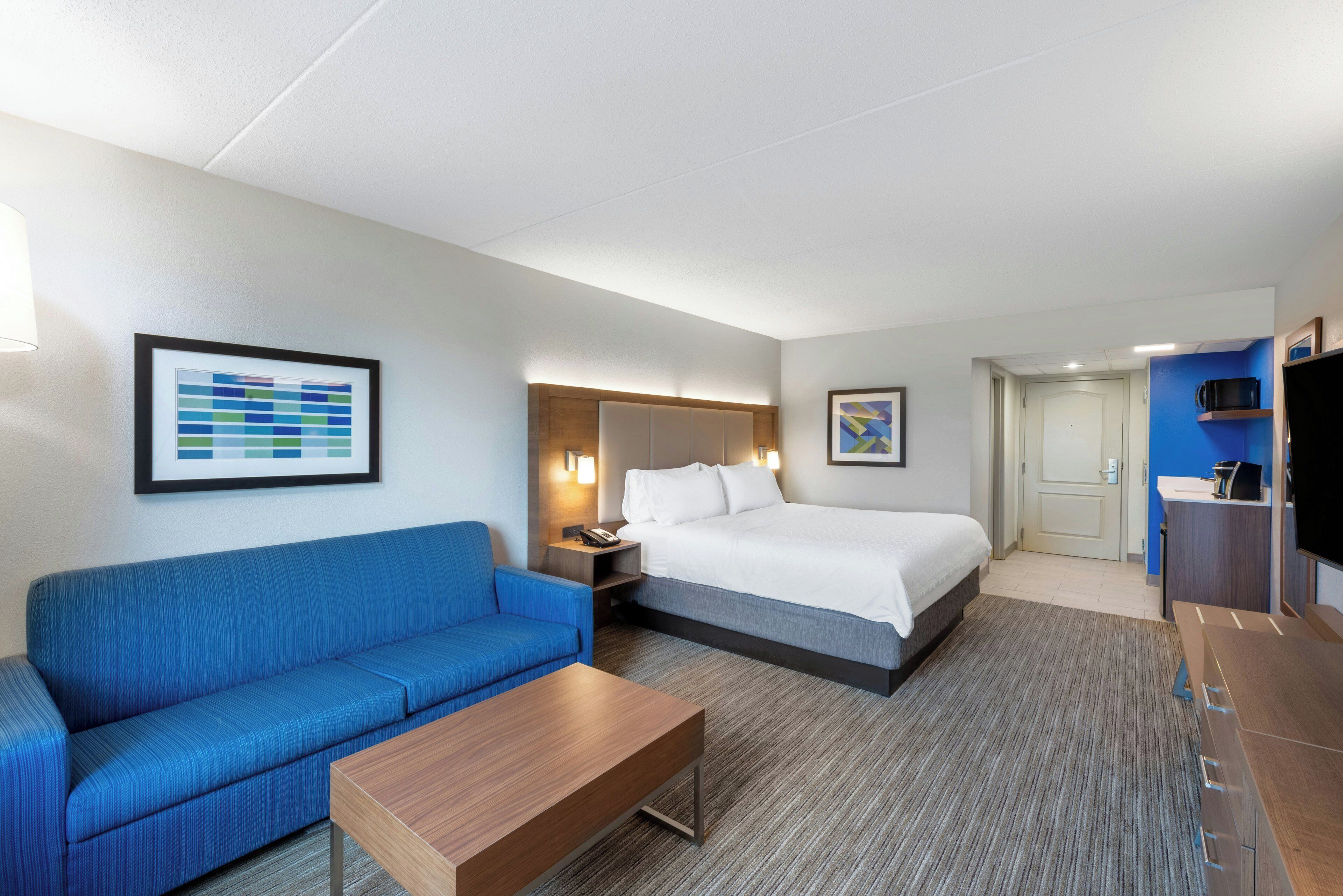 A hotel room with a bed, a blue sofa, a wooden coffee table, a nightstand with a lamp, and a television.