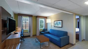 A hotel room with a flat-screen TV, a desk with a chair, a blue sofa, a coffee table, and a window with curtains.