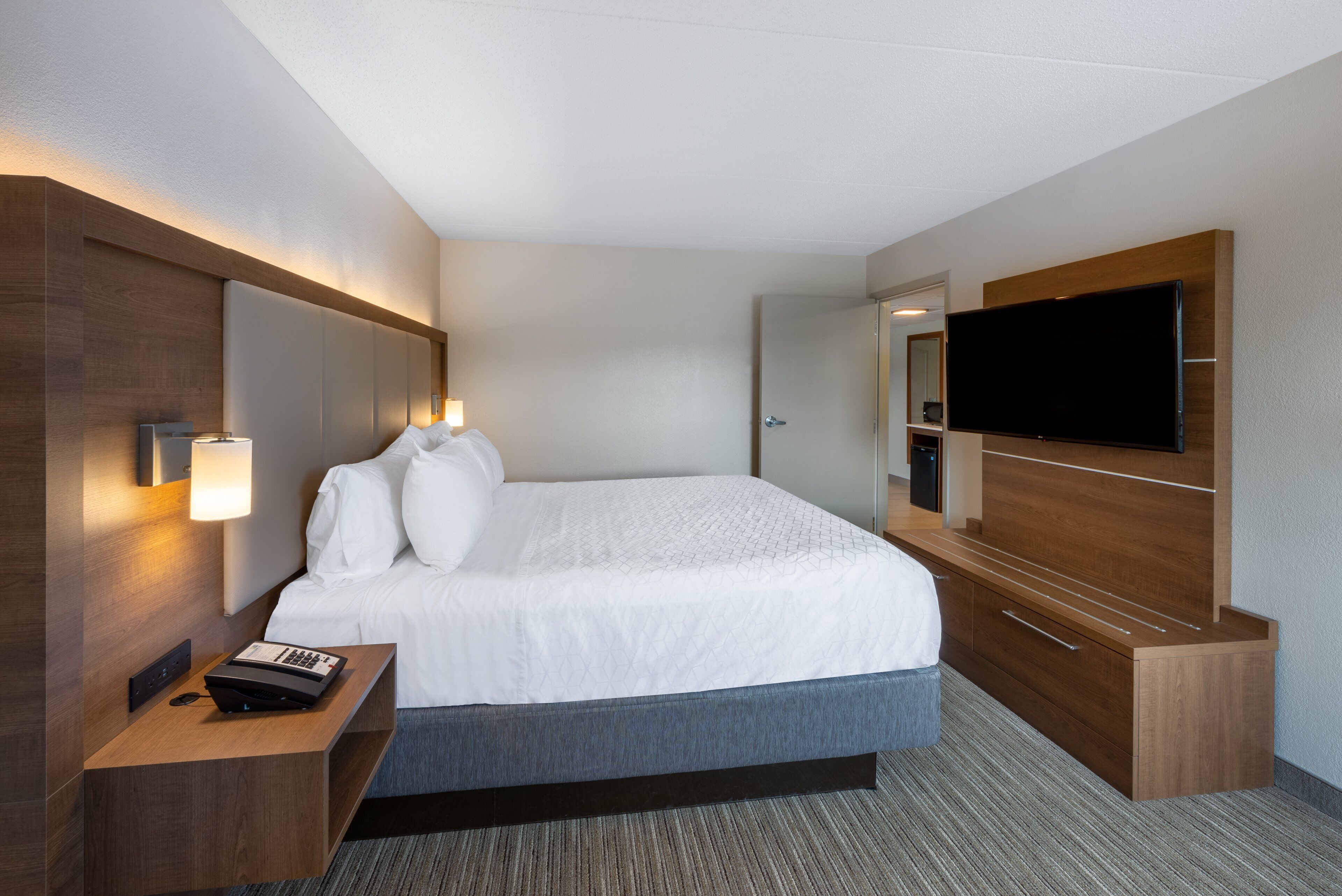 A modern hotel room with a large bed, a desk with a telephone, and a flat-screen TV mounted on the wall.