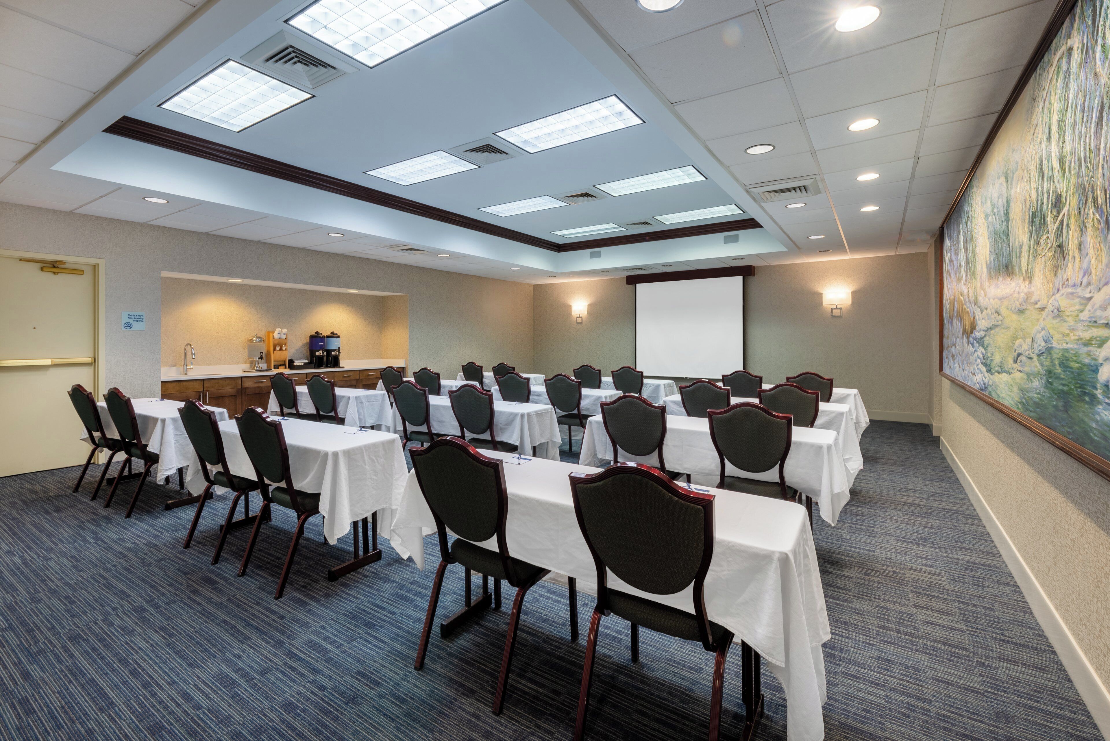A conference room with multiple tables and chairs, a projector screen, and a painting on the wall.
