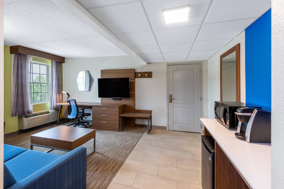 A modern hotel room with a flat-screen TV, a desk, a chair, a sofa, a microwave, a coffee maker, and a door.