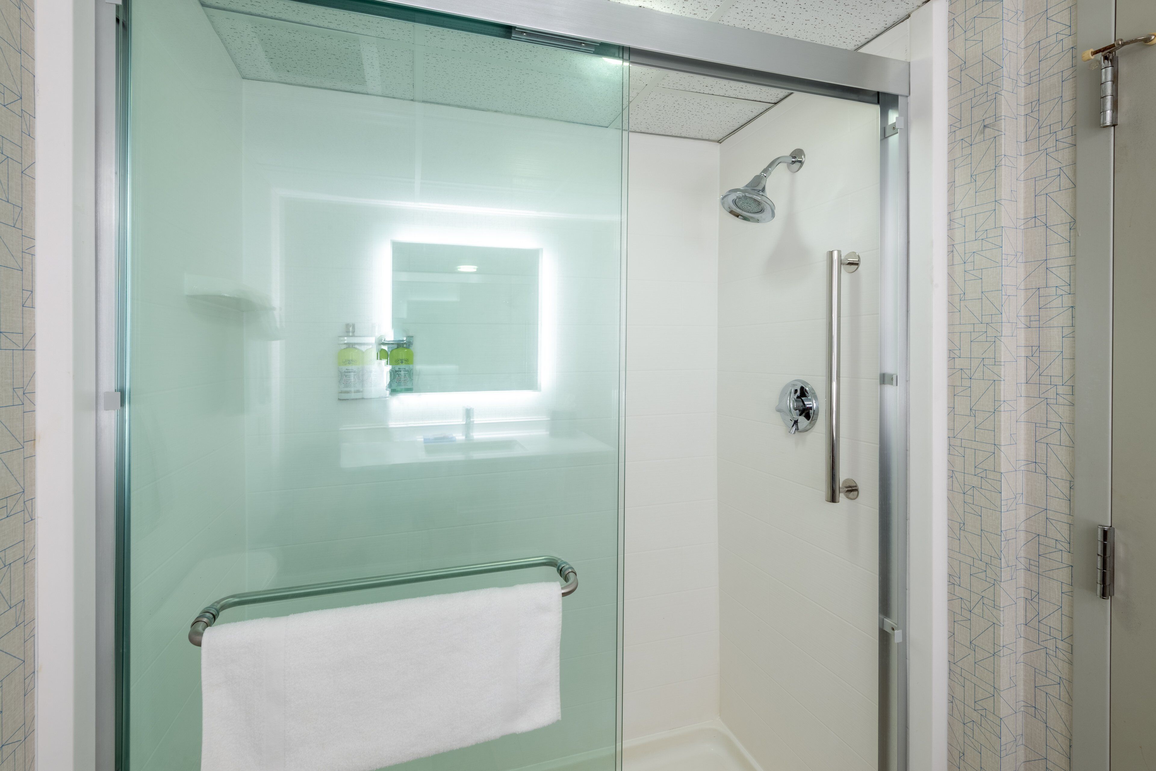 Una ducha con paredes de vidrio y puerta de cristal, una toalla blanca colgada en un perchero metálico y un cabezal de ducha montado en la pared.
