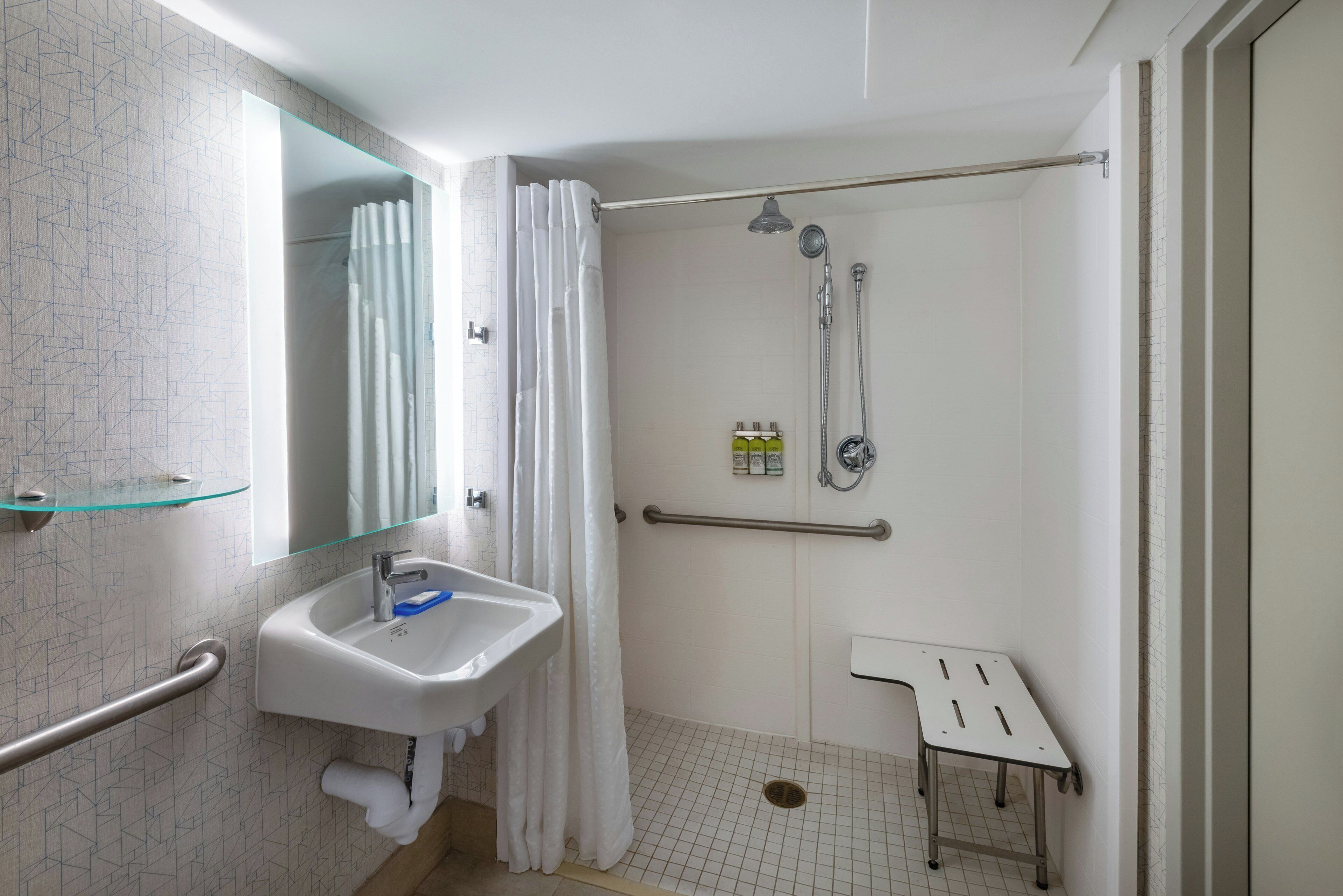 Une salle de bain moderne avec une étagère en verre, un lavabo, une douche avec un pommeau de douche à main et une barre d’appui.