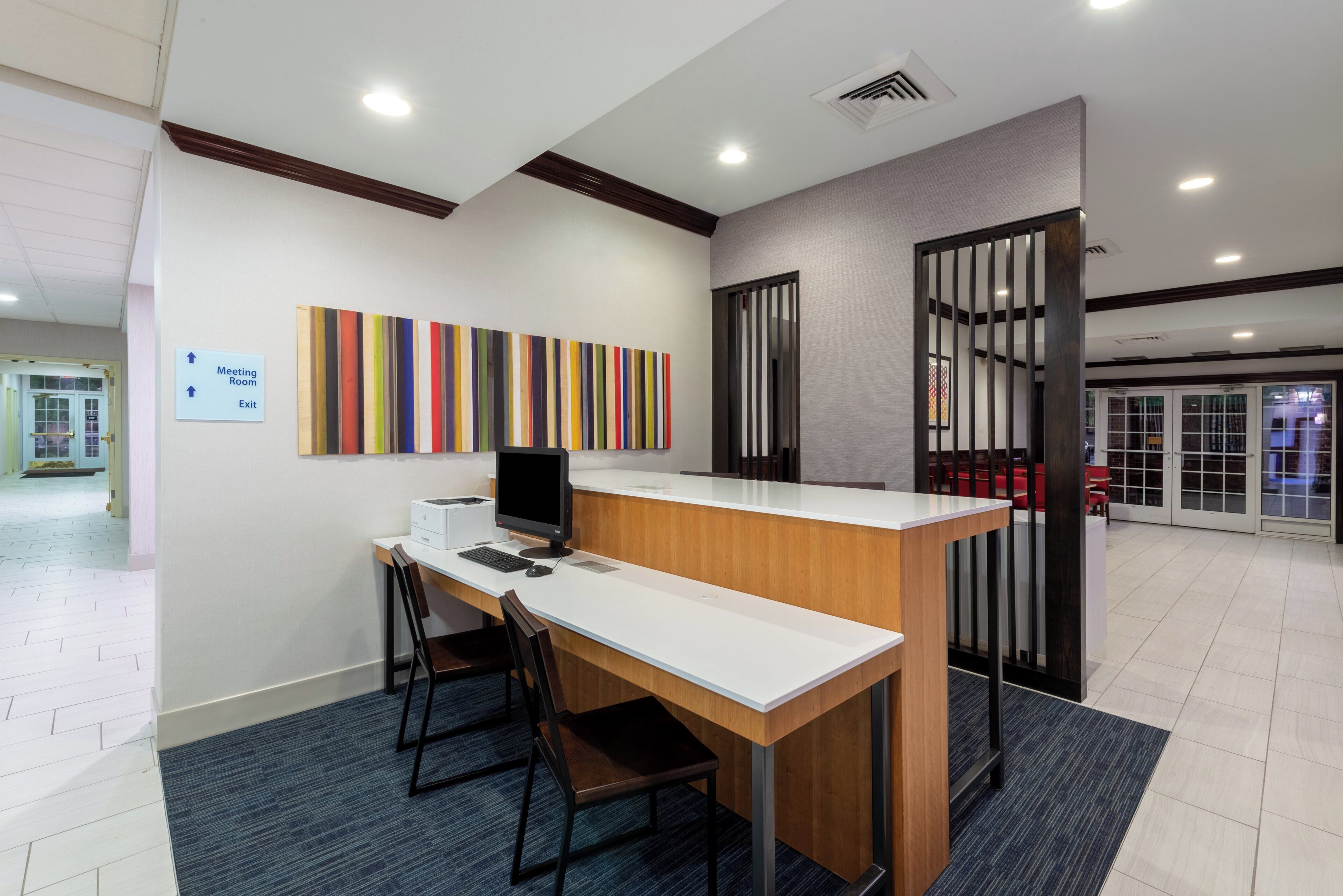 A reception desk with a computer, two chairs, a colorful artwork on the wall, and a sign indicating the meeting room and exit.