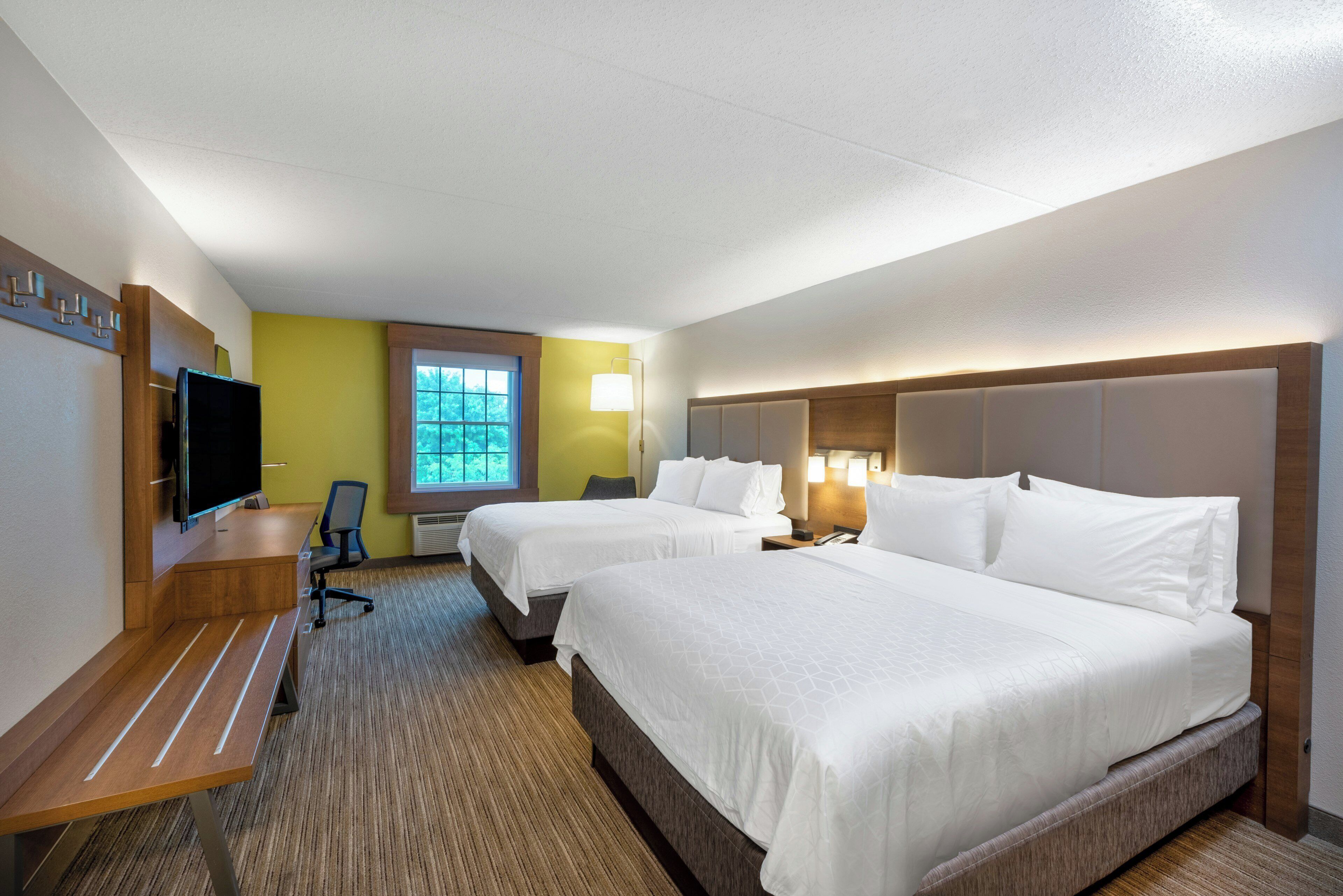 A hotel room with two beds, a desk, a chair, a television, and a window with a view of trees.