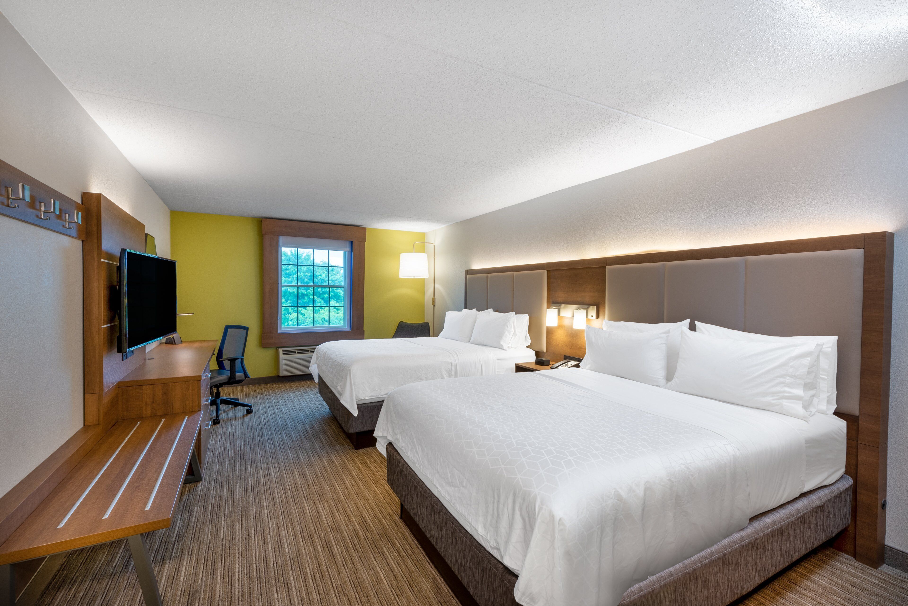 A hotel room with two beds, a desk, a television, and a window with a view of trees.