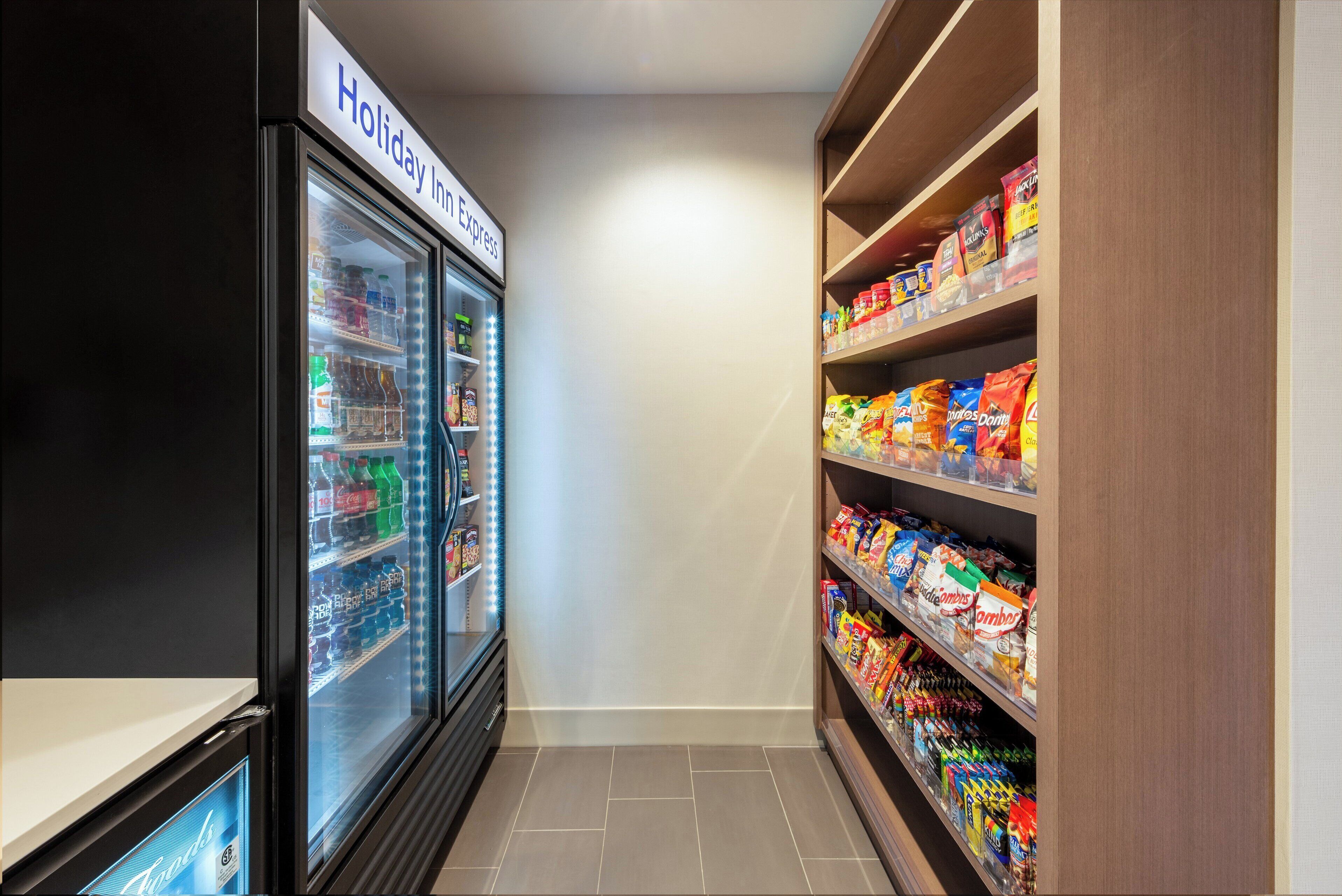 A narrow hallway with a Holiday Inn Express refrigerator stocked with beverages and snacks, and shelves filled with various packaged goods.