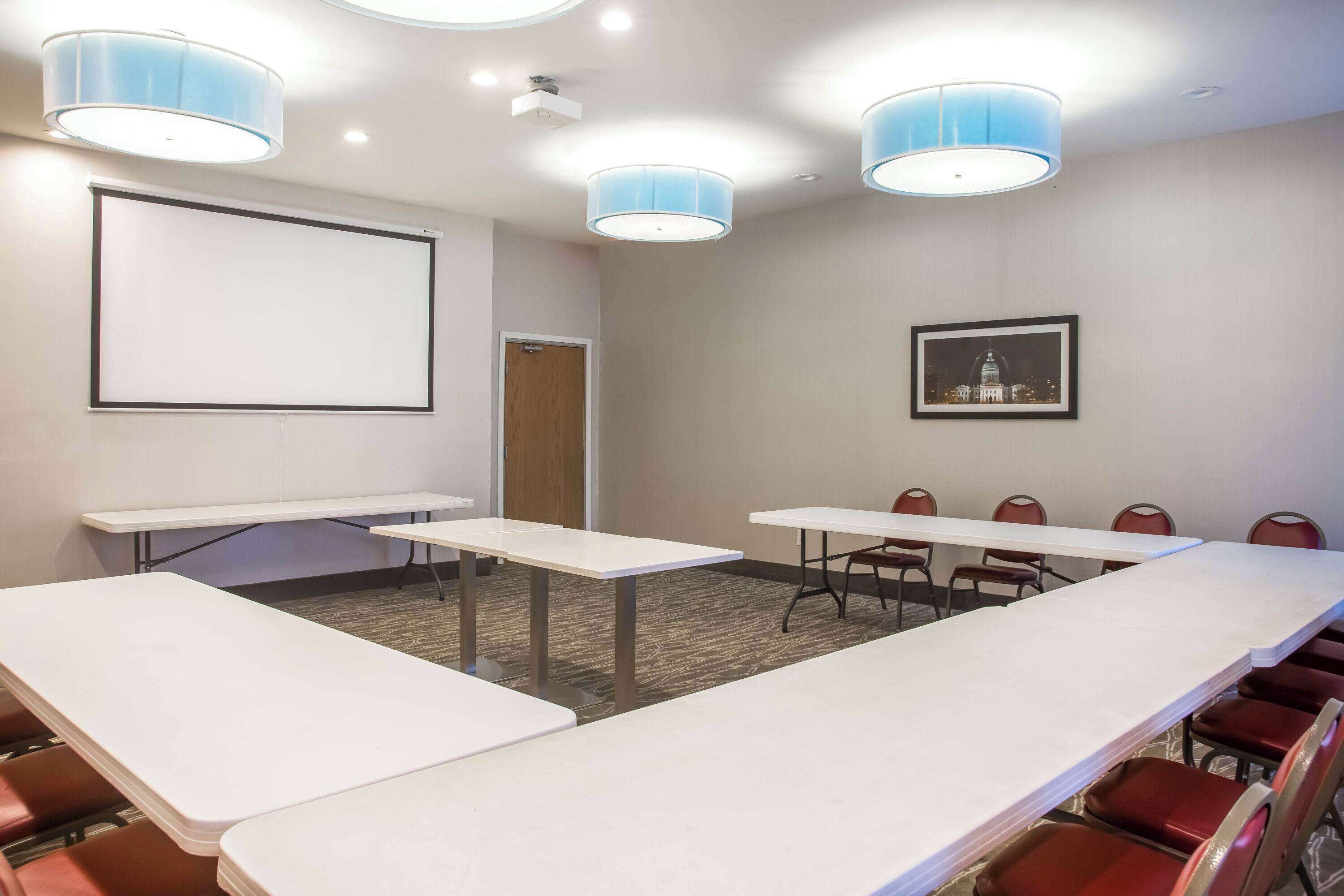 Un salón de conferencias con una gran mesa, sillas, una pantalla de proyector e iluminación en el techo.