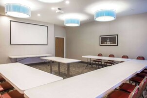 Un salón de conferencias con una gran mesa, sillas, una pantalla de proyector e iluminación en el techo.
