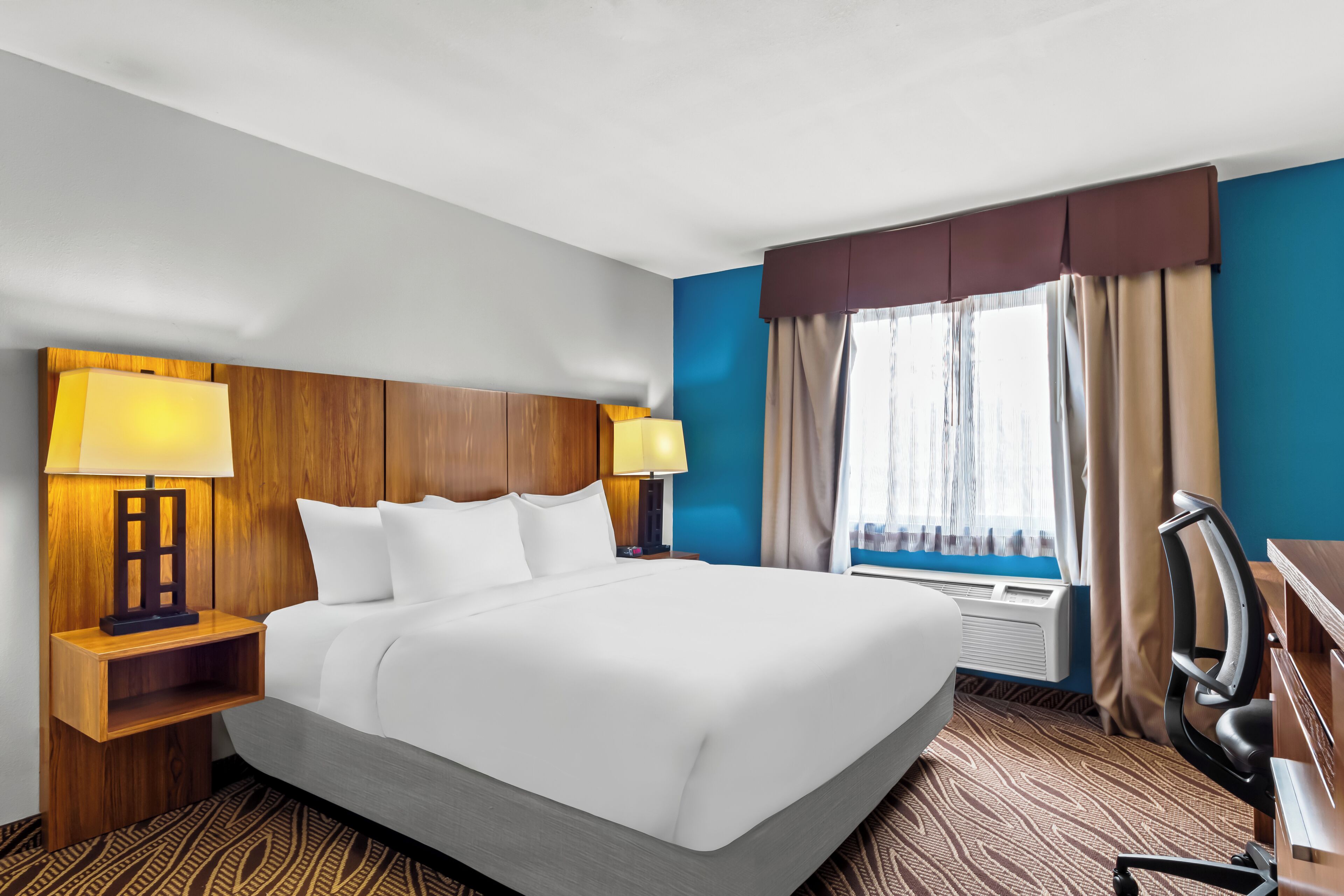 A hotel room with a large bed, two bedside lamps, a desk with a chair, a window with curtains, and a wall heater.