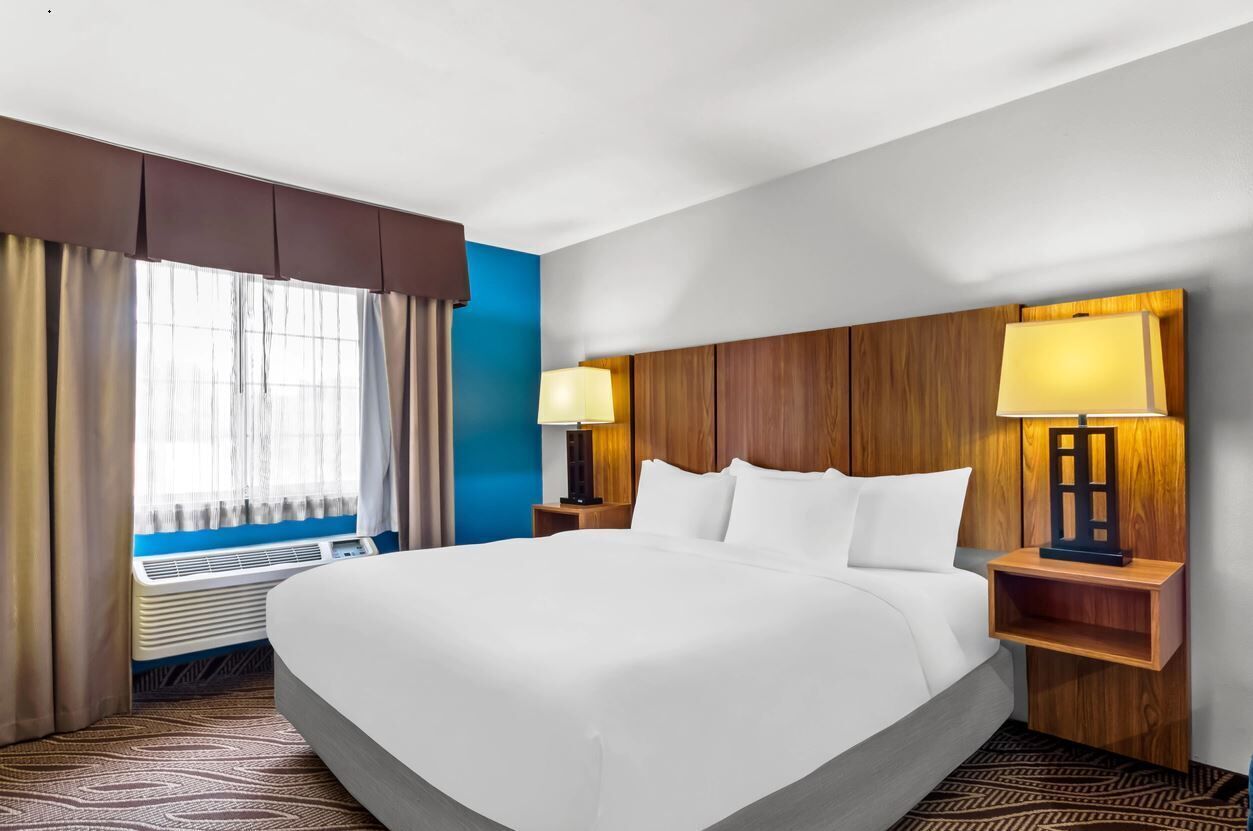 A hotel room with a large bed, two bedside lamps, a window with curtains, and a wall-mounted heater.