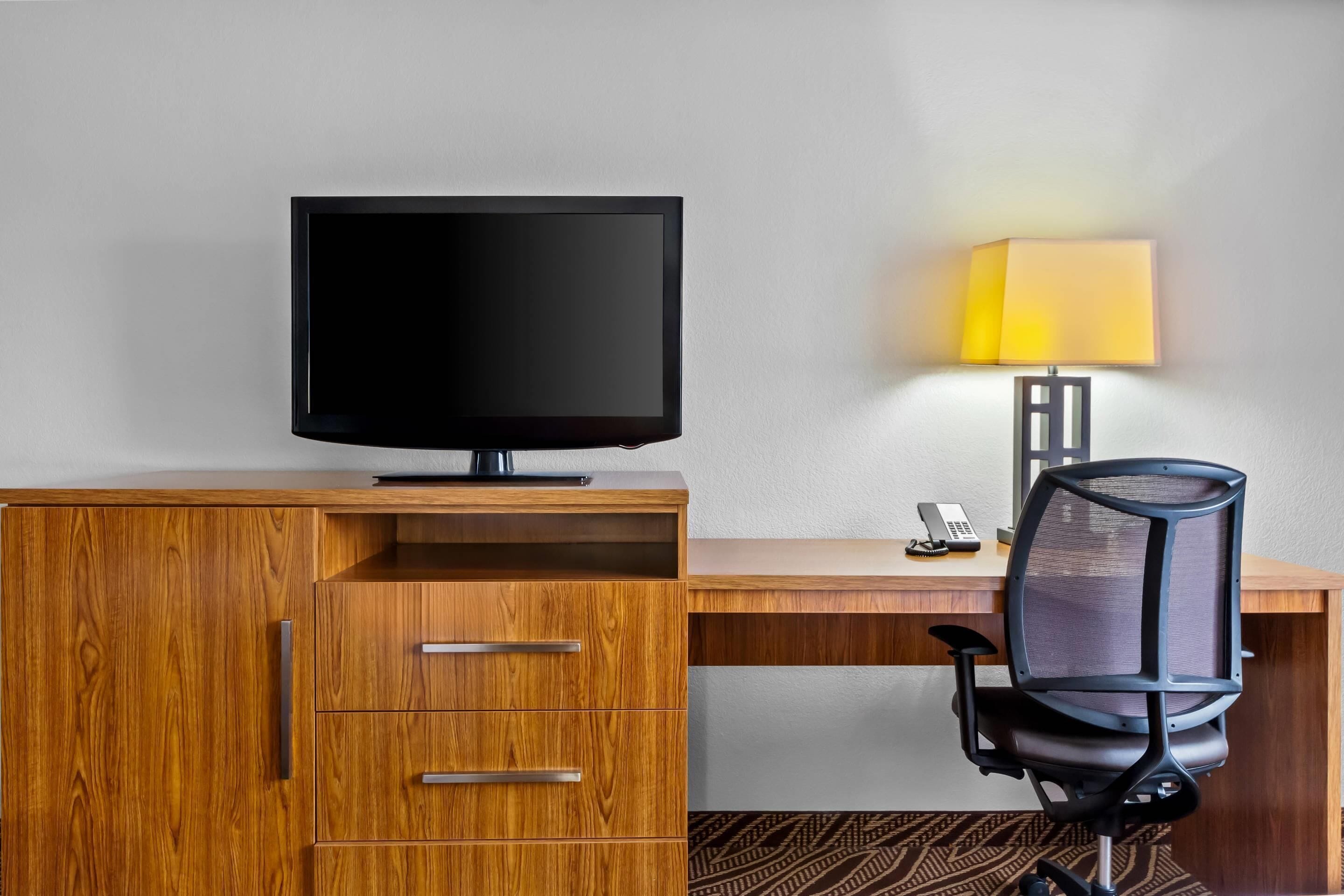 A hotel room with a wooden desk, a television, a chair, and a lamp.