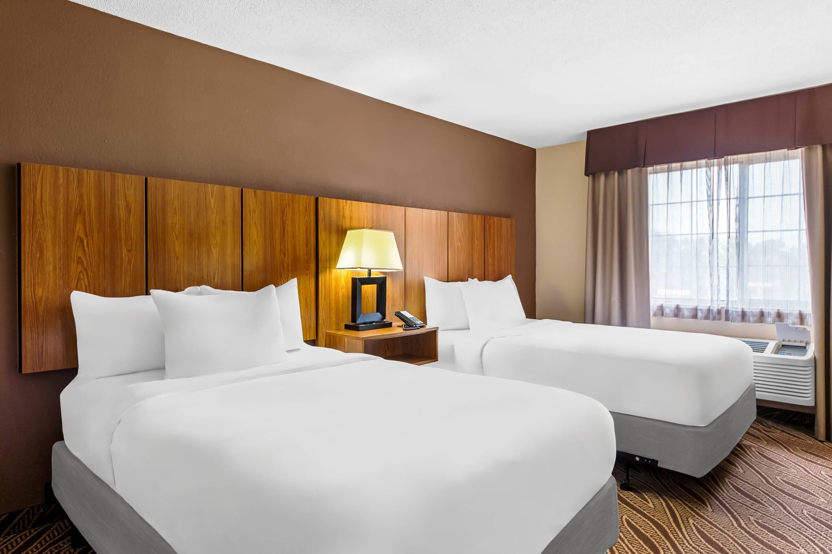 Habitación de hotel con dos camas, cabecero de madera, mesita de noche con lámpara y ventana con cortinas.